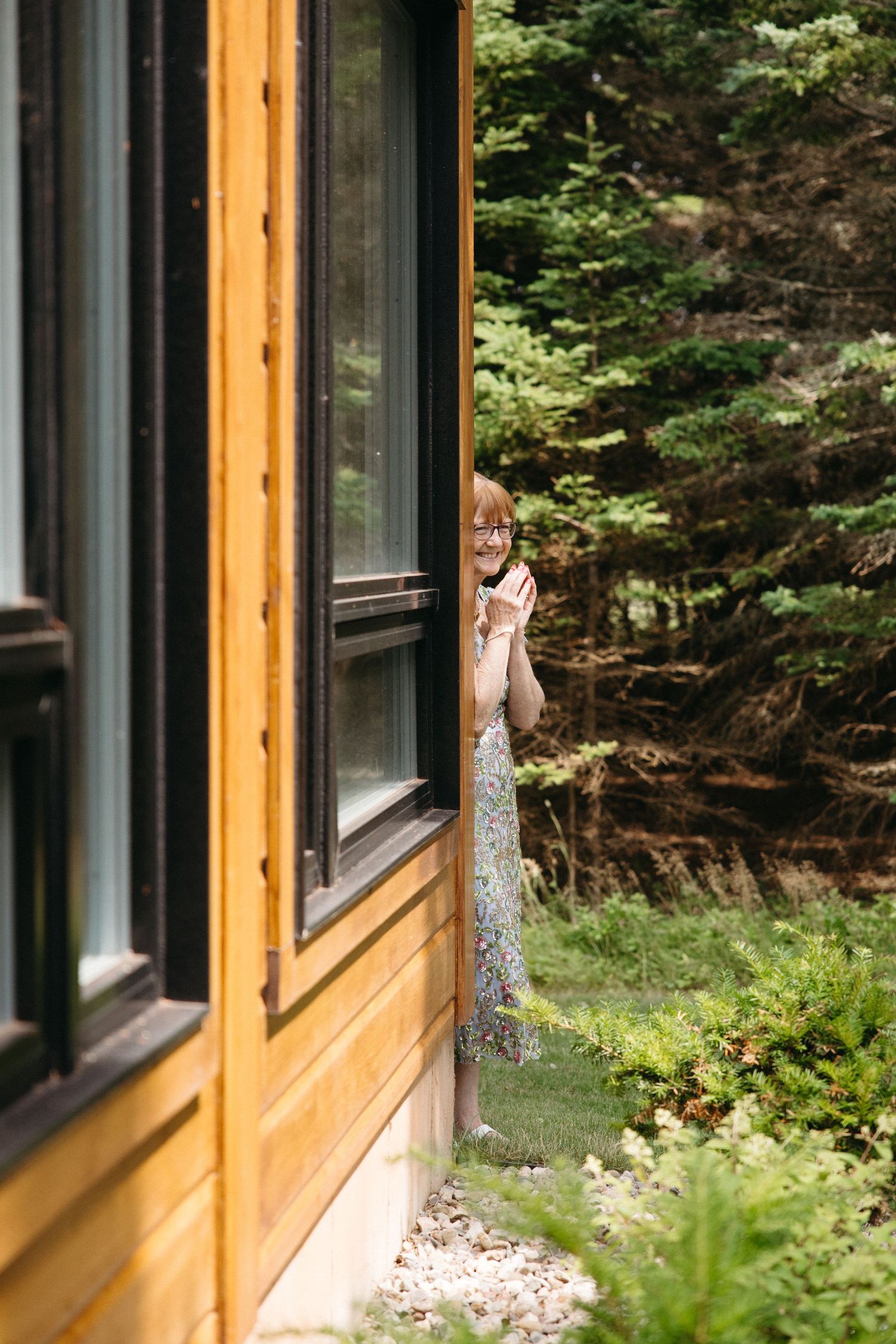 Woman in sequined dress peeking around corner of wooden cabin surrounded by lush green forest.