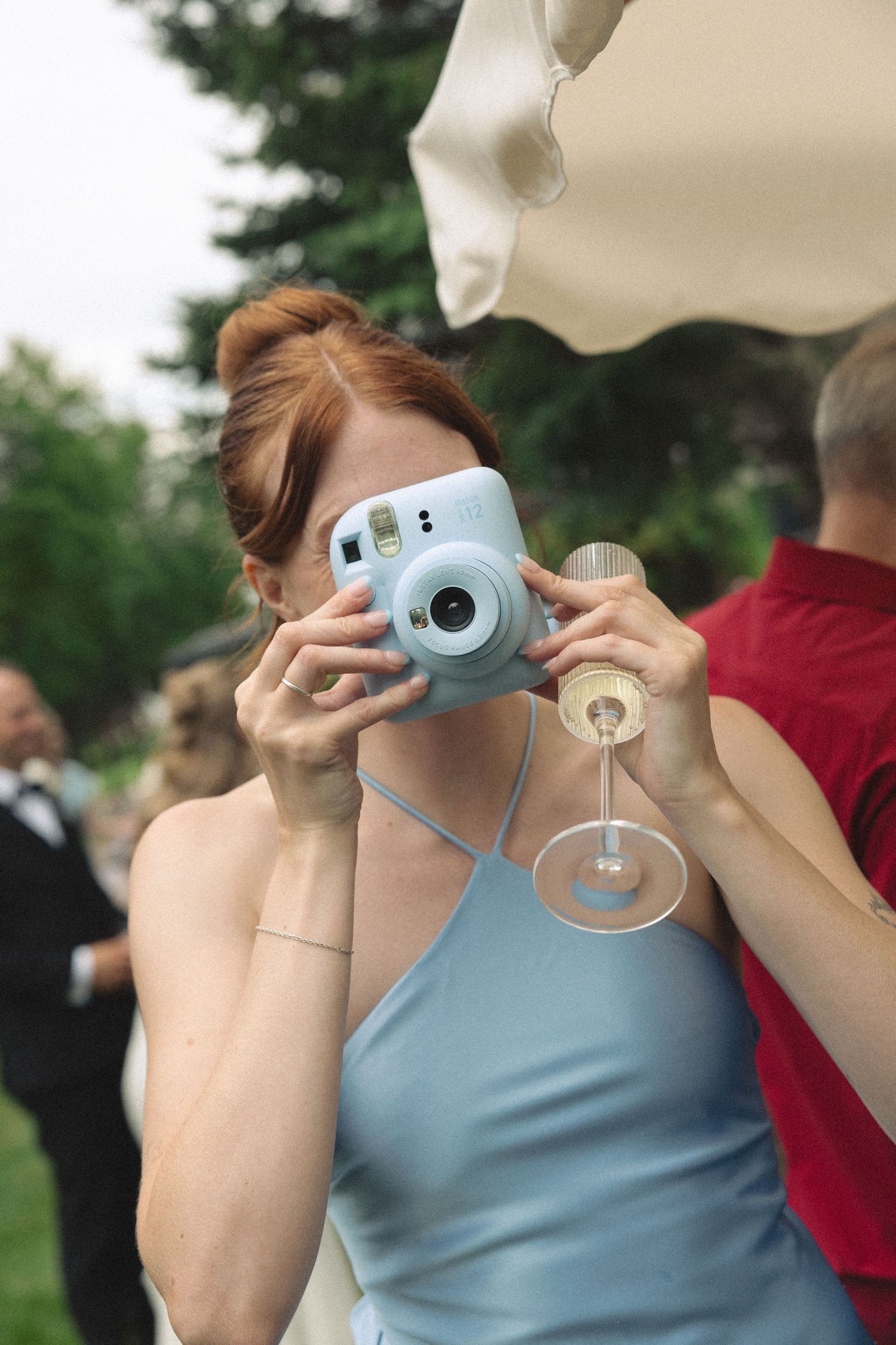 Red-haired bridesmaid in blue dress photographs with a Instax mini camera while holding champagne.
