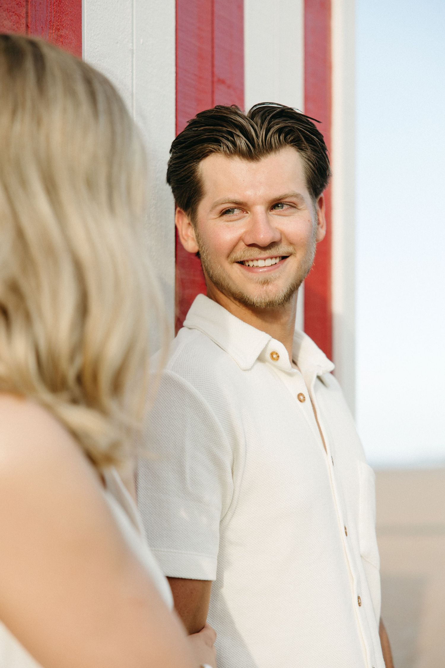 Smiling man in white shirt leaning against red striped wall at beach.