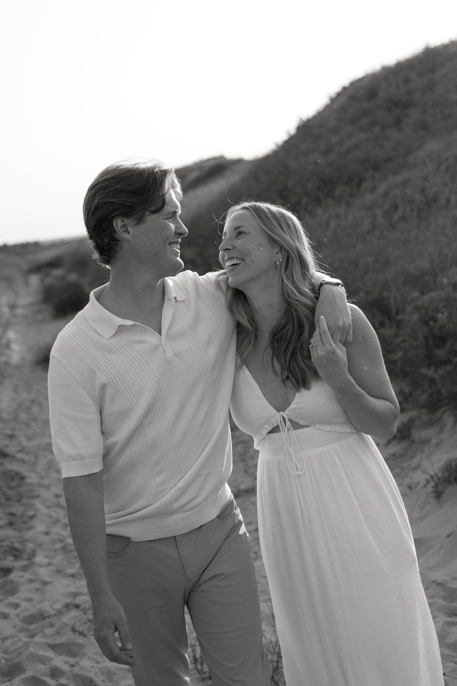 Black and white photo of laughing couple walking together along sandy beach dunes.