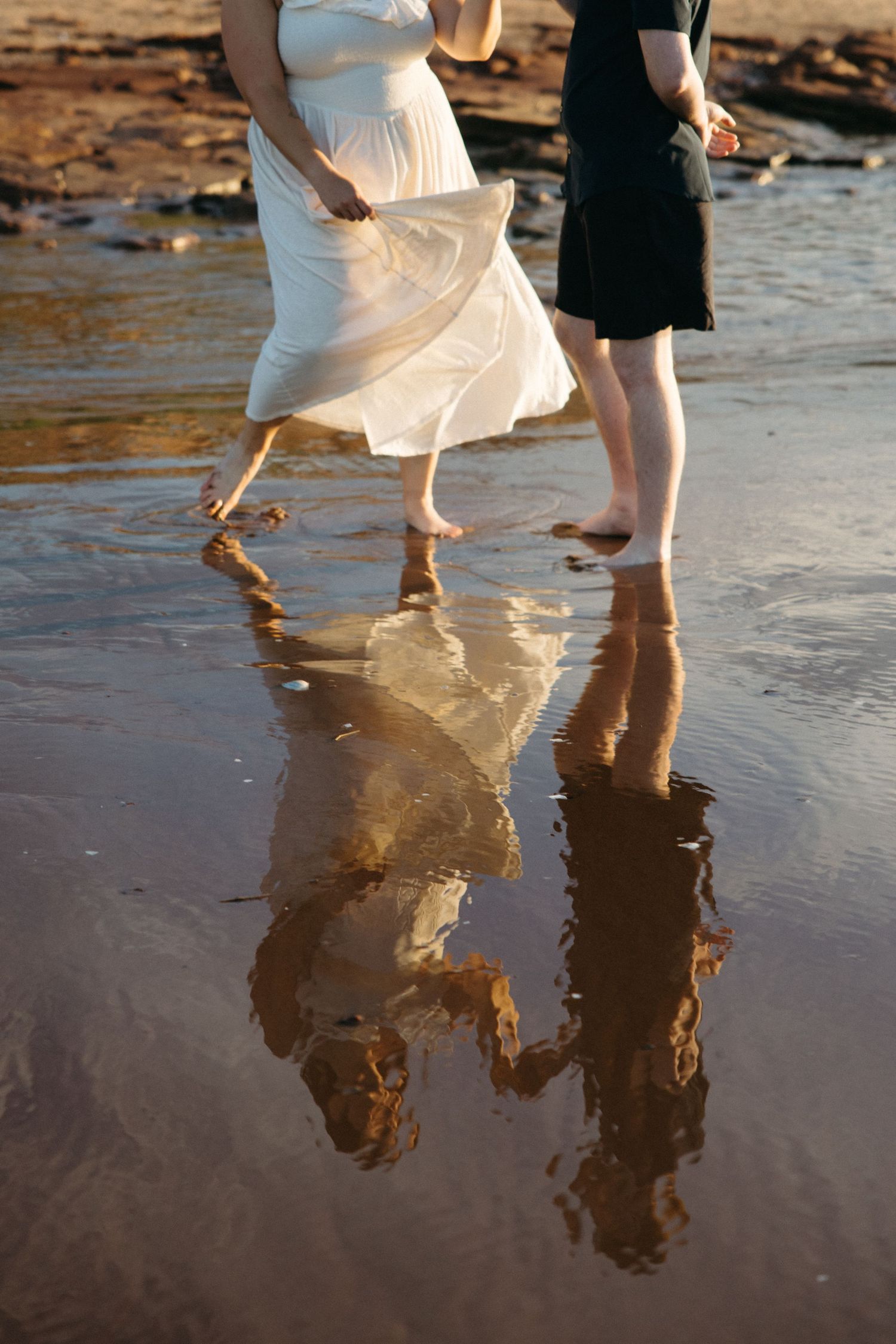 Couple walking barefoot on wet sandy beach, white dress flowing, reflections visible in shallow water at golden hour.