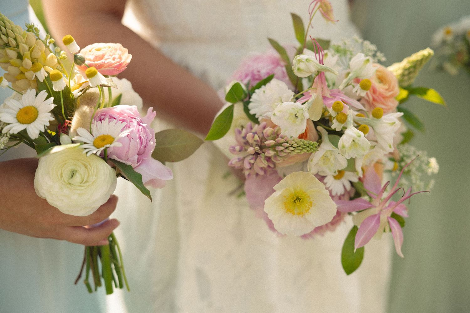 Two brides holding pastel wedding bouquets with peonies, daisies, and ranunculus in pink, white, and yellow hues.