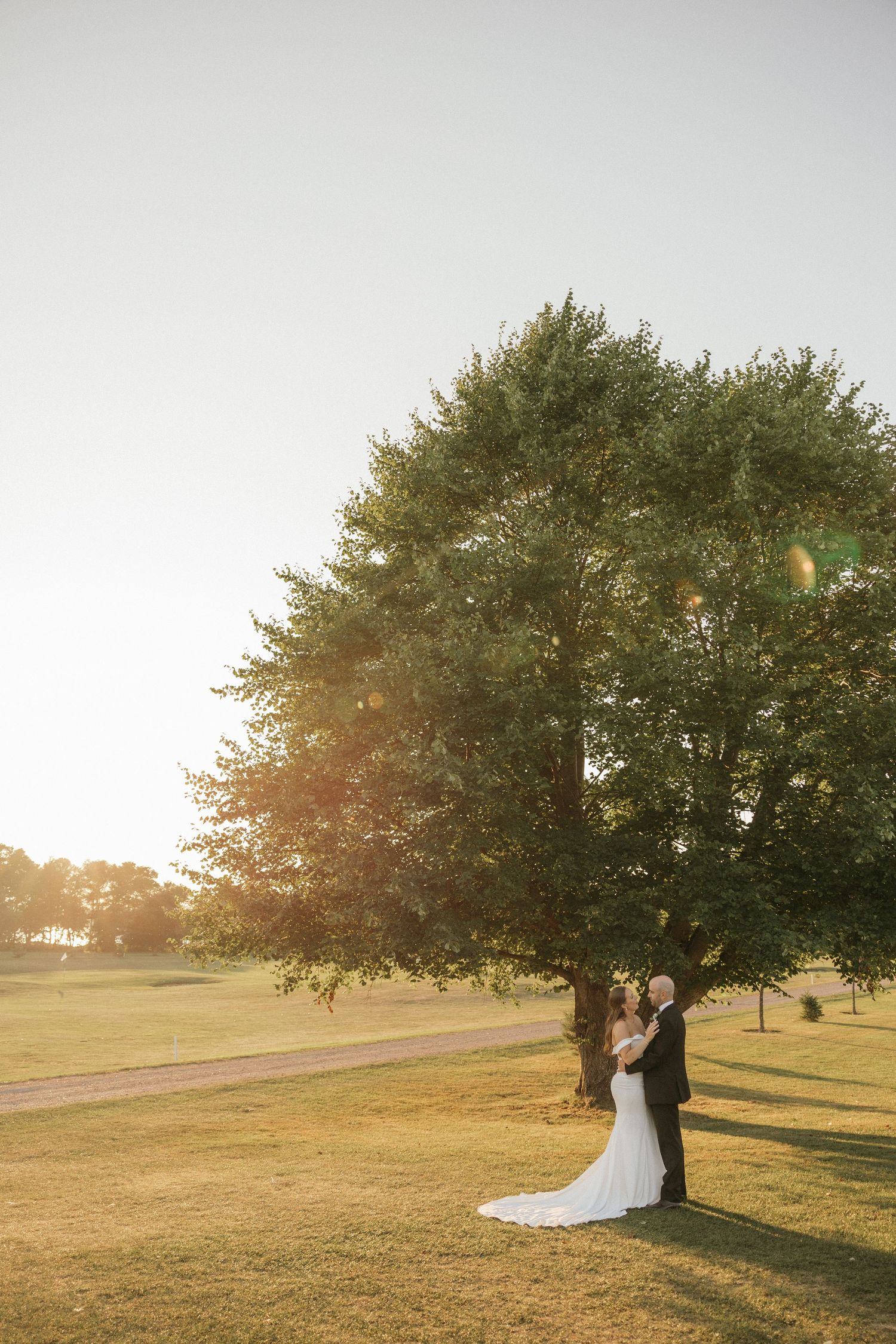 A couple poses for romantic sunset wedding photos under a large tree in a grassy field.