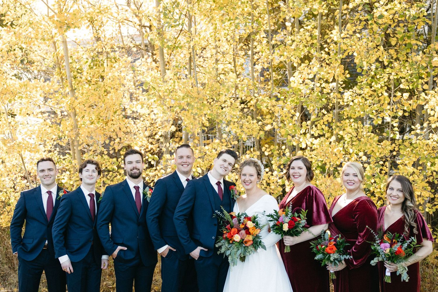 Wedding party in navy suits and burgundy dresses posed against autumn trees.