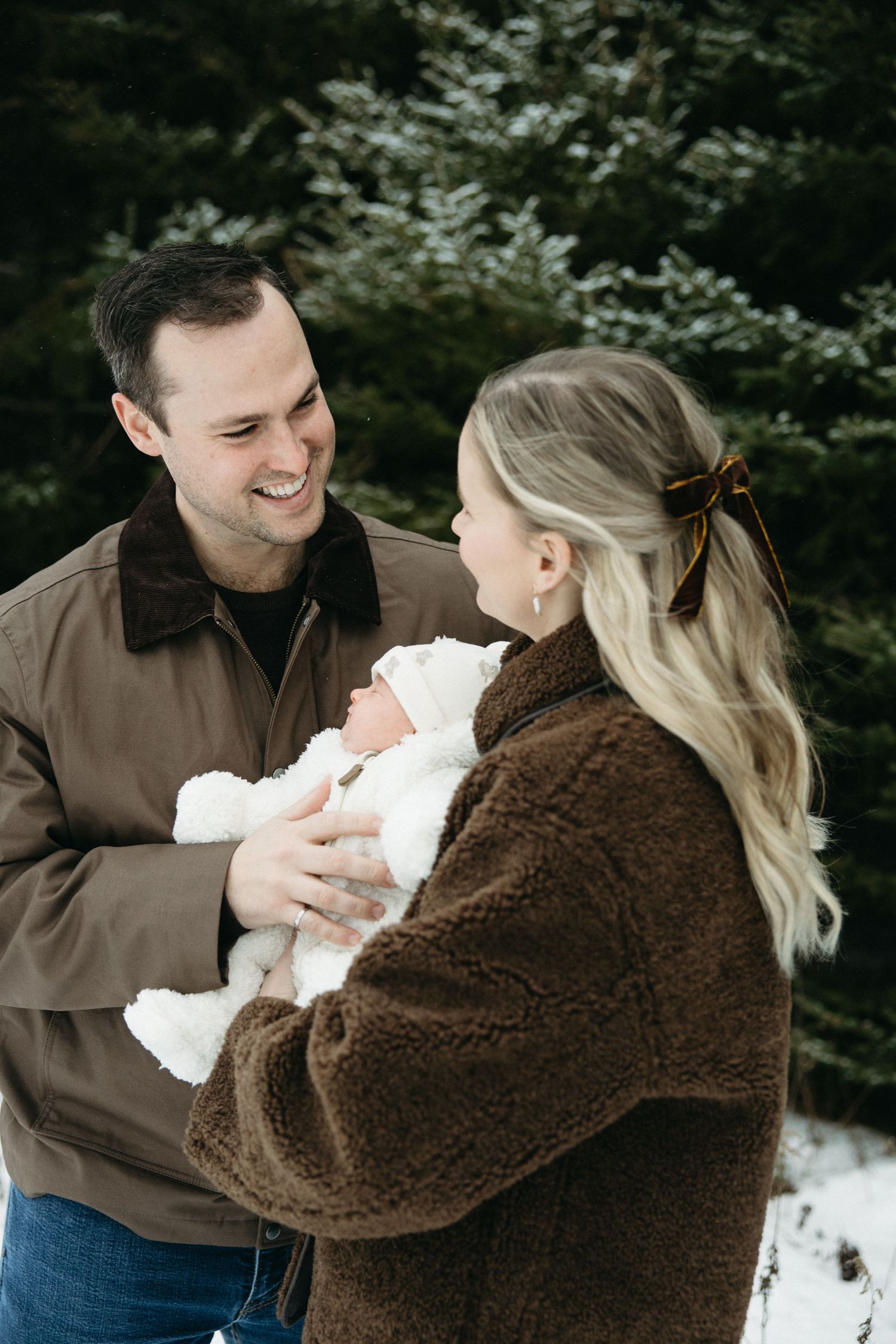 A couple in winter coats share a tender moment with their baby against a snowy evergreen backdrop.
