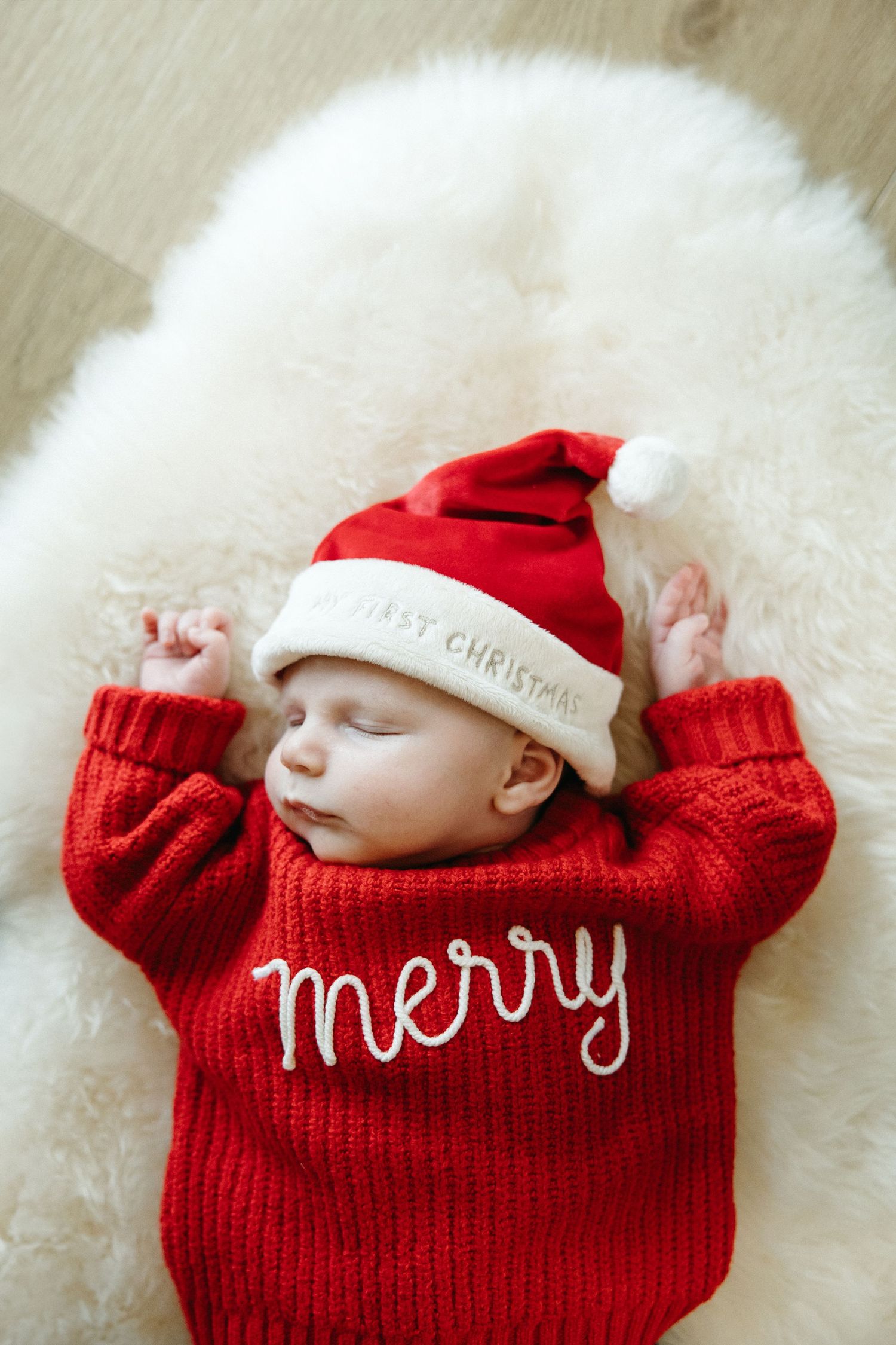 A sleeping infant in festive red 'Merry' sweater and Santa hat lies on plush white carpet.