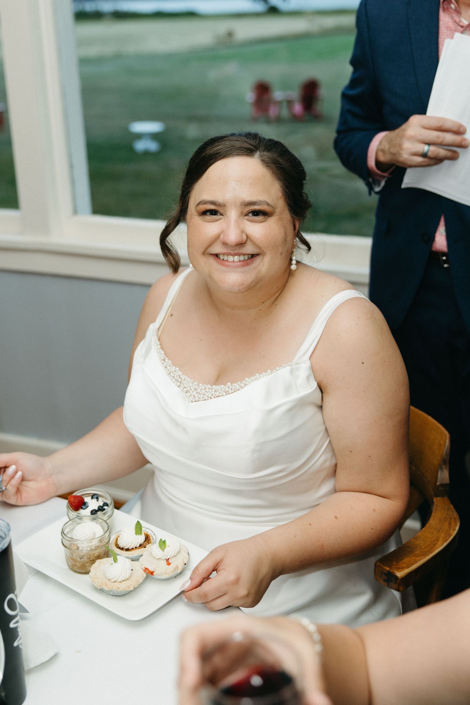 Happy bride in white wedding dress sitting at reception table smiling with desserts in front of her.
