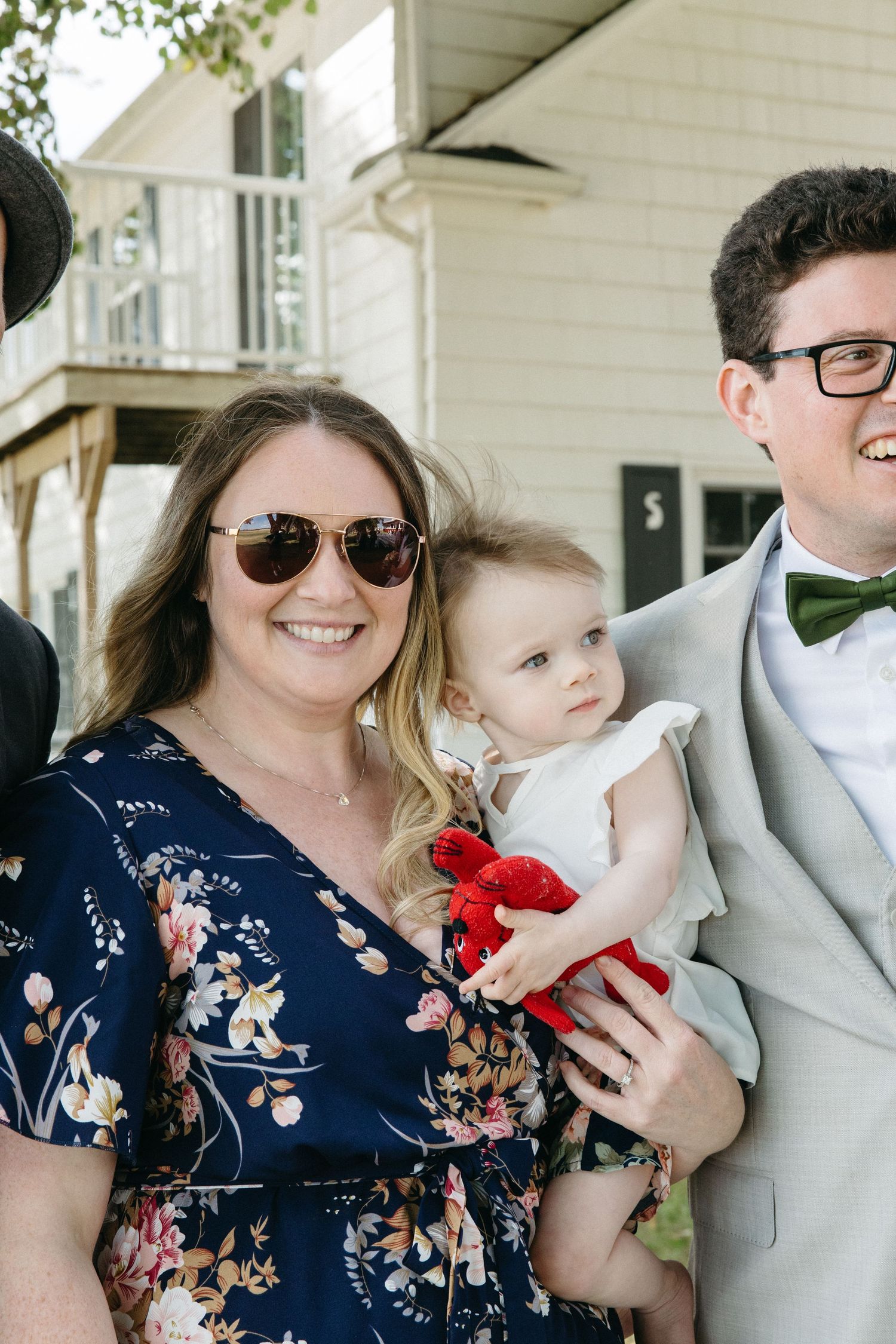 A woman in a floral dress holds a baby in white while a man in a gray suit with green bow tie poses outdoors.