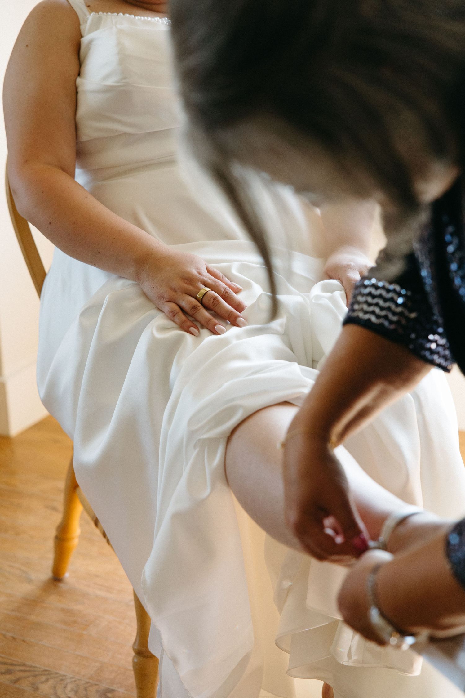 Bride in white gown sits as bridesmaid helps fasten wedding shoe during bridal preparations.
