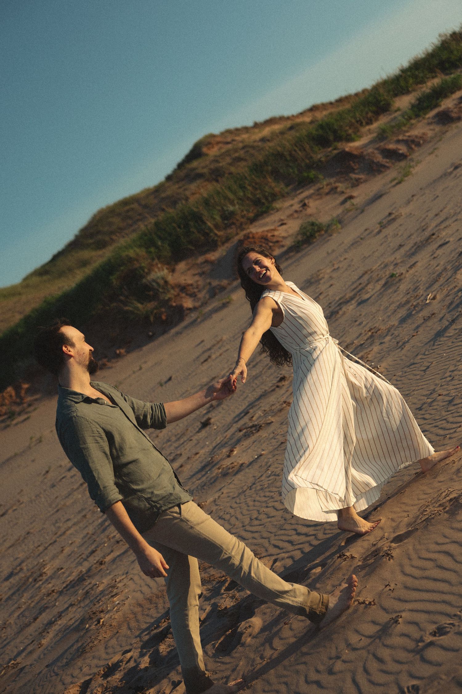 Couple holding hands on sandy dunes, woman in flowing white striped dress leaning back joyfully.