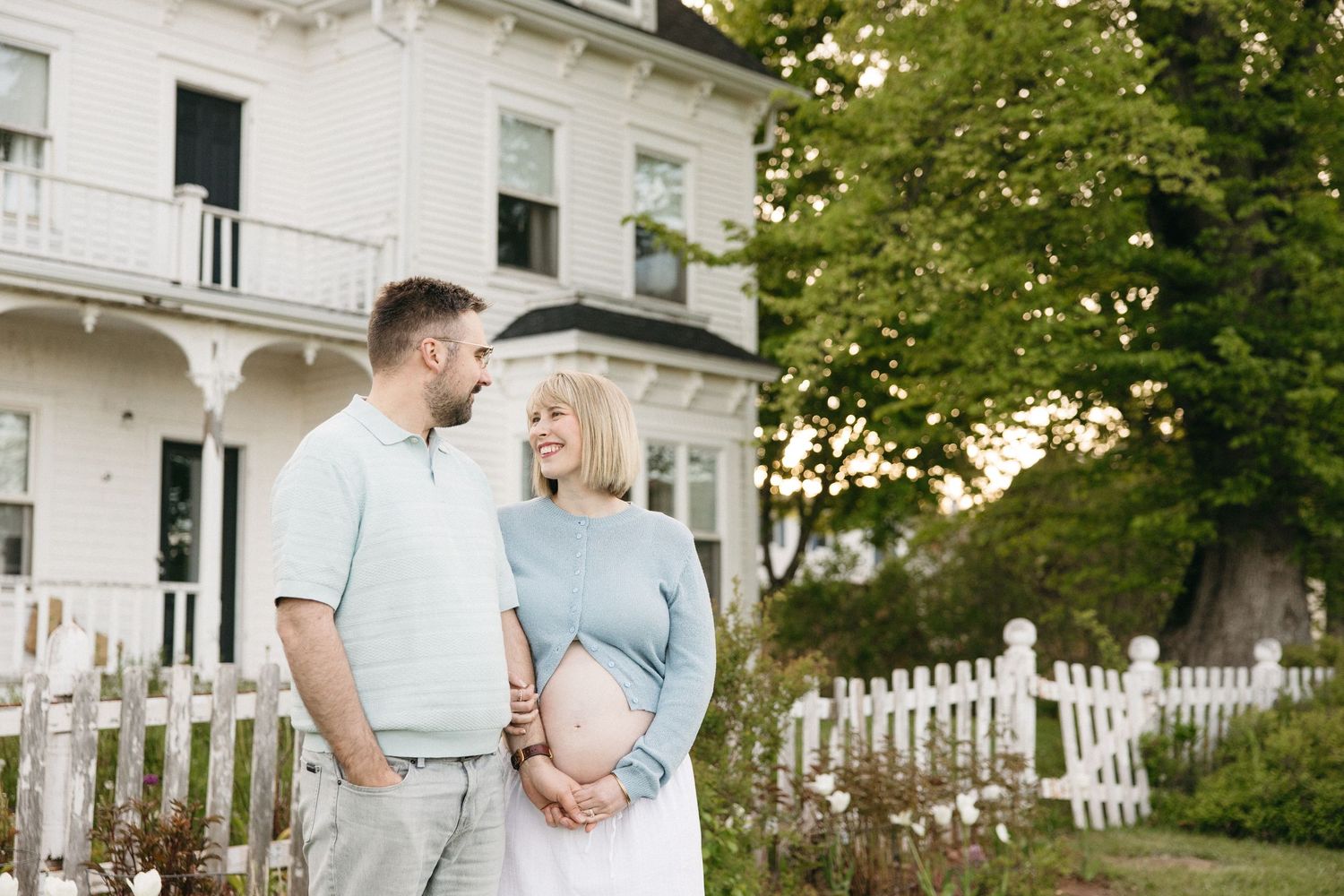 Pregnant couple smiling at each other in front of a white Victorian house with a picket fence.