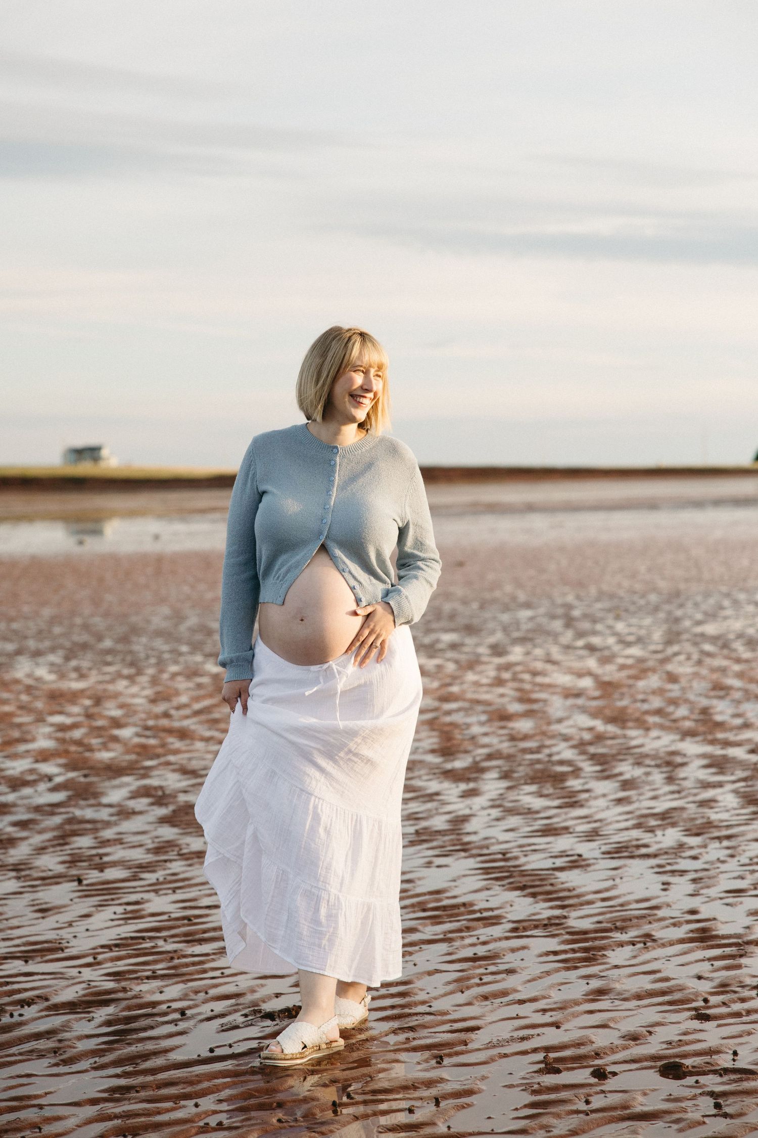 Smiling pregnant woman in blue top and white skirt cradling her baby bump on a sandy beach at golden hour.