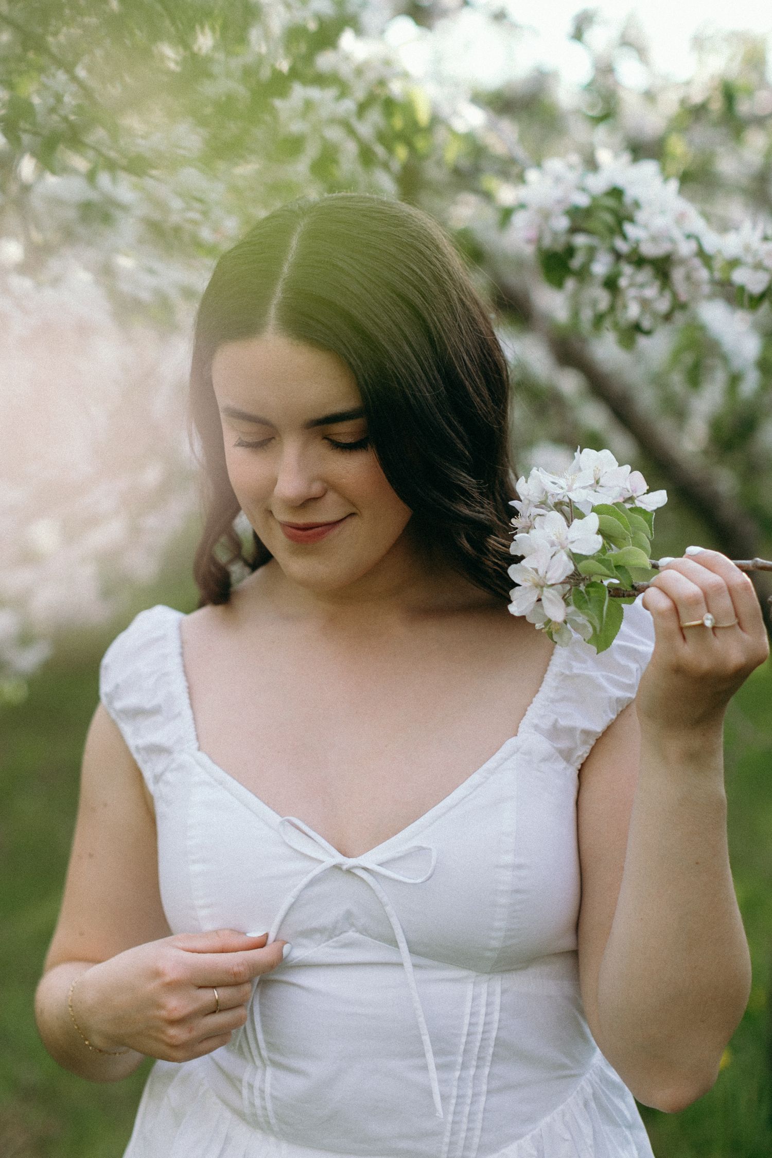 Young woman in white dress holding white spring blossoms, smiling softly in a blooming garden.