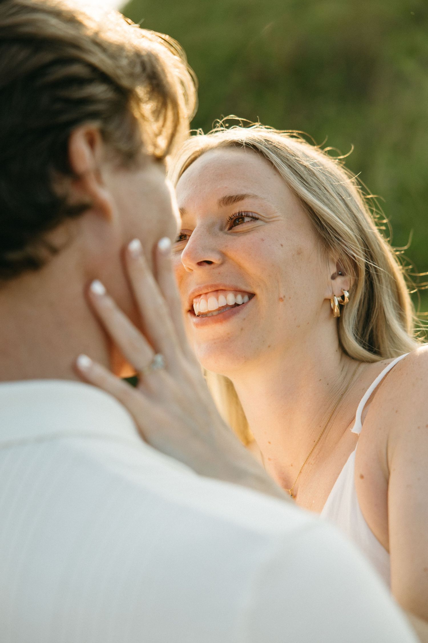 Close-up of smiling blonde woman with freckles touching mans face in warm golden sunlight outdoors.