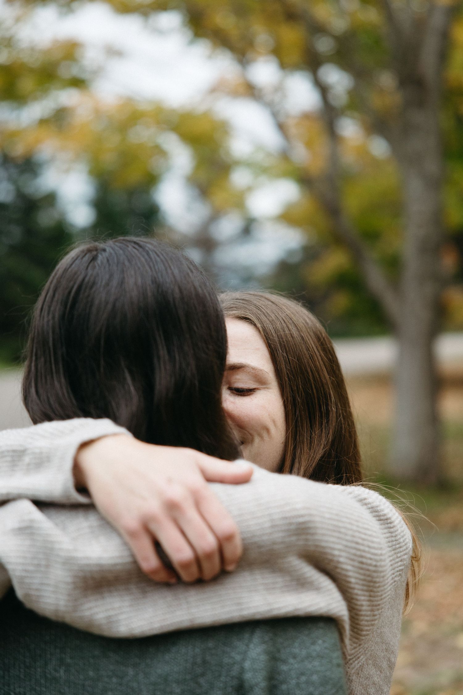 Two women embrace in a warm hug, one smiling with closed eyes, with blurred autumn trees in the background.
