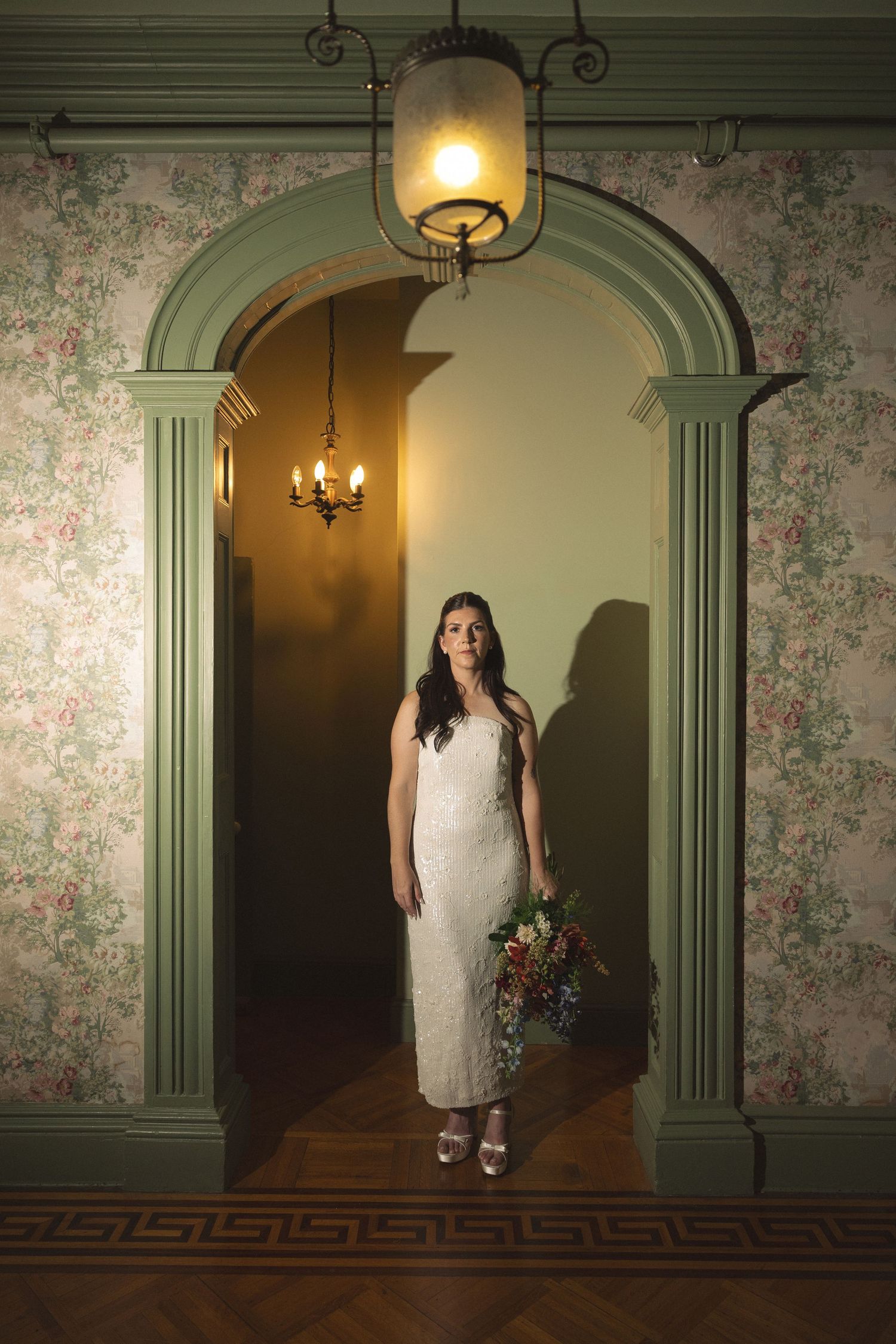 A bride in a white dress holds a colorful bouquet while standing in a green arched doorway with floral wallpaper and a hanging lantern.