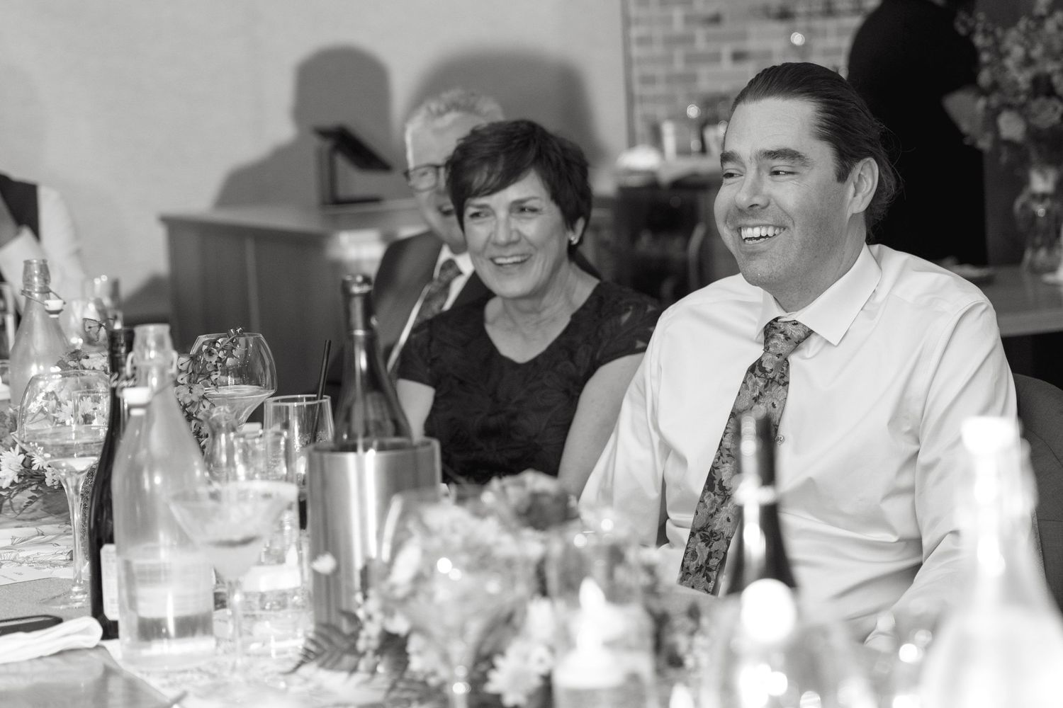 Black and white photo of a smiling man and woman seated at a reception table with wine bottles, glasses and flowers.