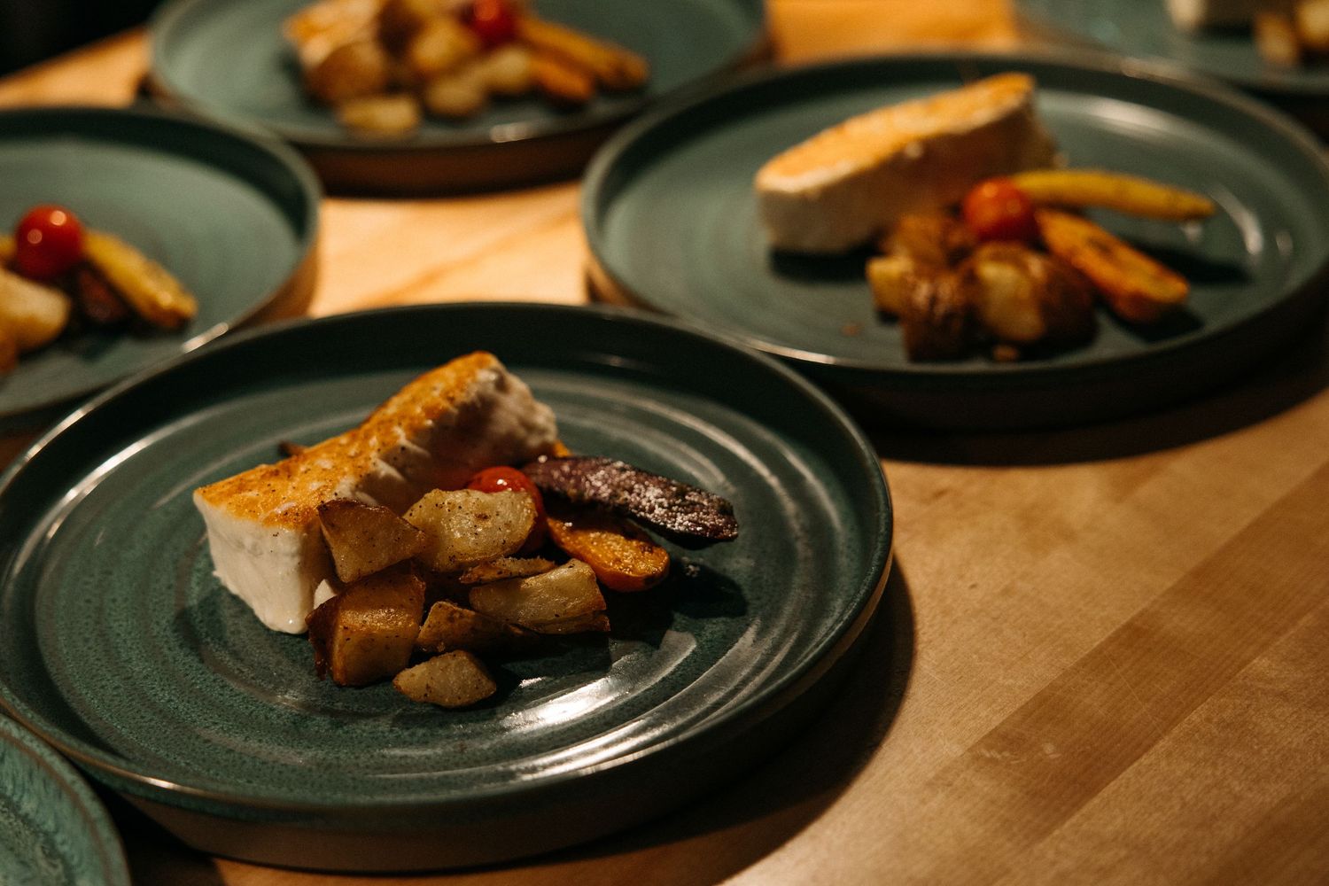 Plated fish fillets with roasted root vegetables and cherry tomatoes served on dark green ceramic plates arranged on a rustic wooden table.
