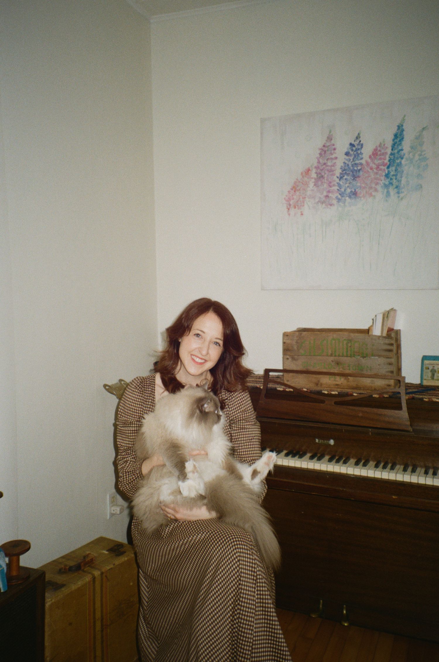 A smiling woman holds a fluffy Ragdoll cat while seated in front of a vintage upright piano indoors.