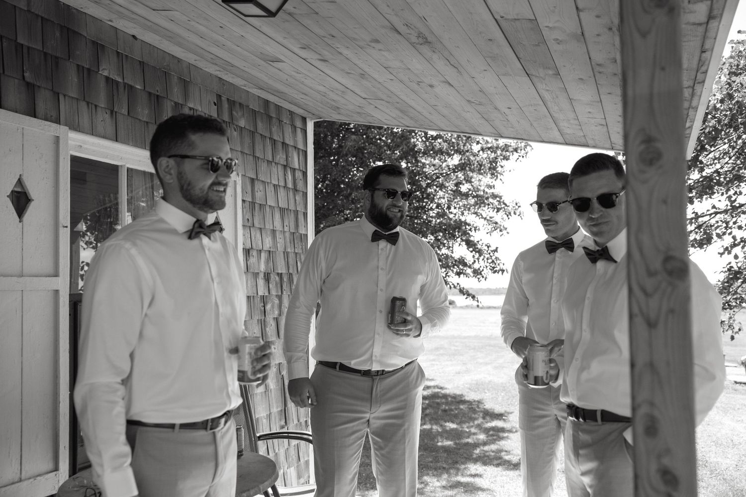 Black and white photo of three groomsmen in white shirts and bow ties relaxing on a porch holding drinks.