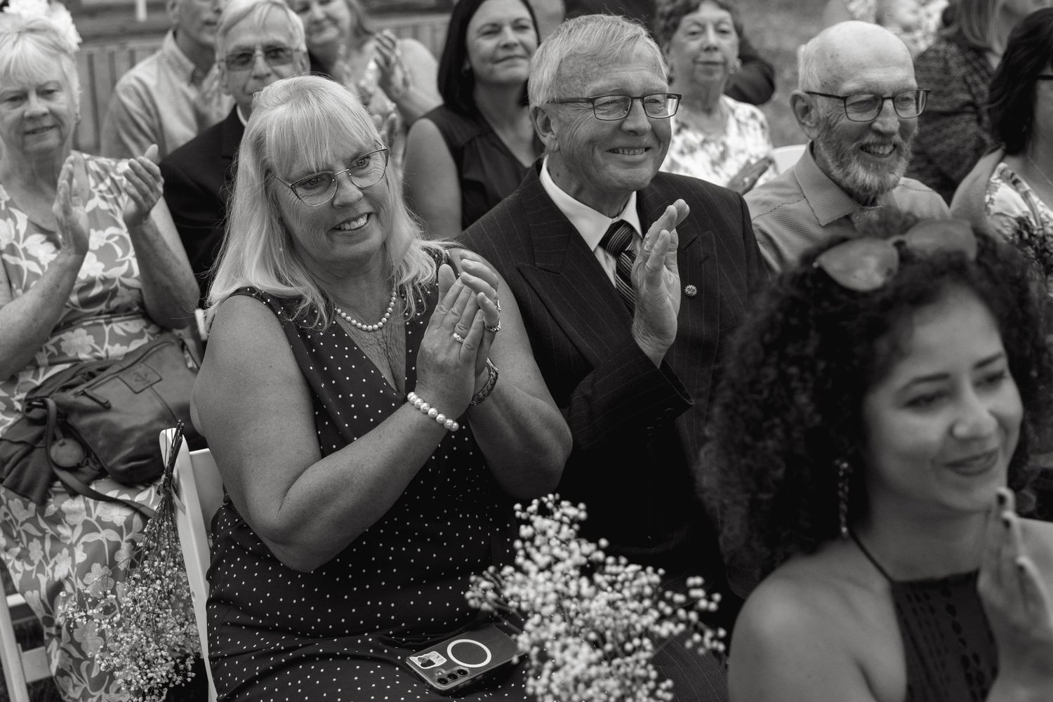 Joyful wedding guests clap and smile during an outdoor ceremony in this black and white photo.