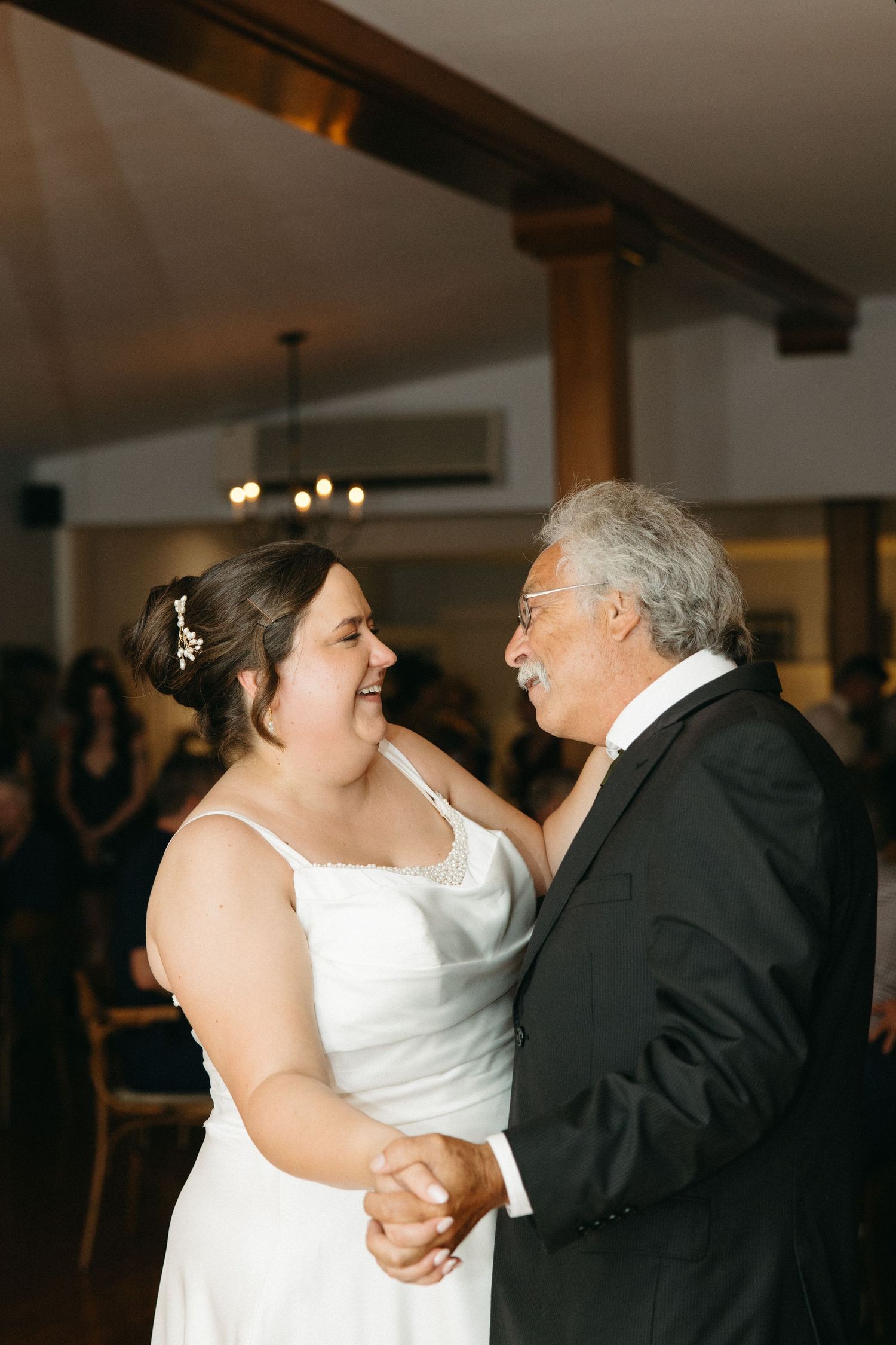 Bride shares joyful father-daughter dance at indoor wedding reception with wooden beam ceiling.