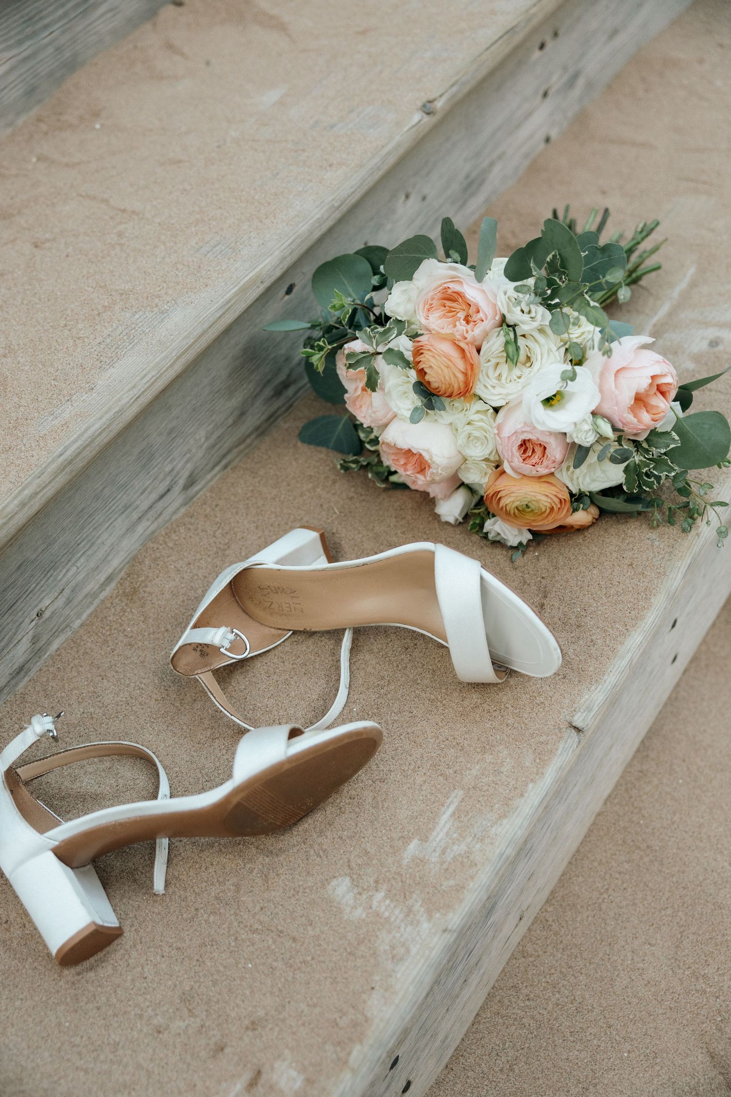 White bridal heels and peach rose bouquet resting on wooden steps.