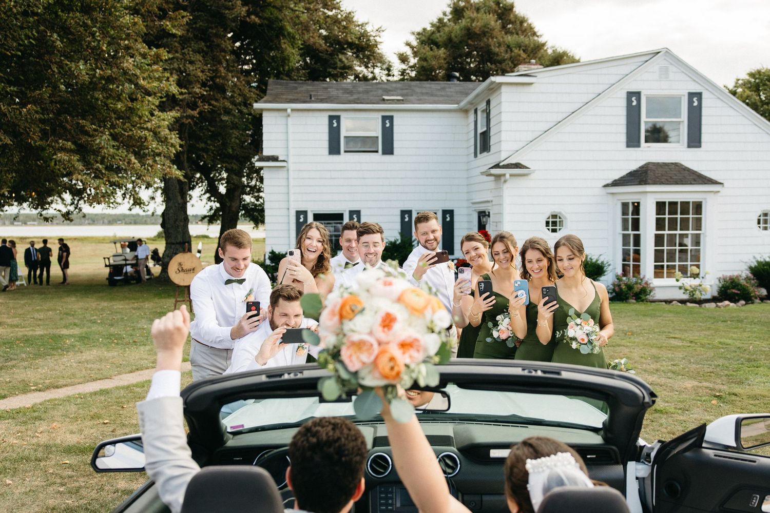 Excited wedding party cheers as bride and groom depart in a convertible car outside a white house.