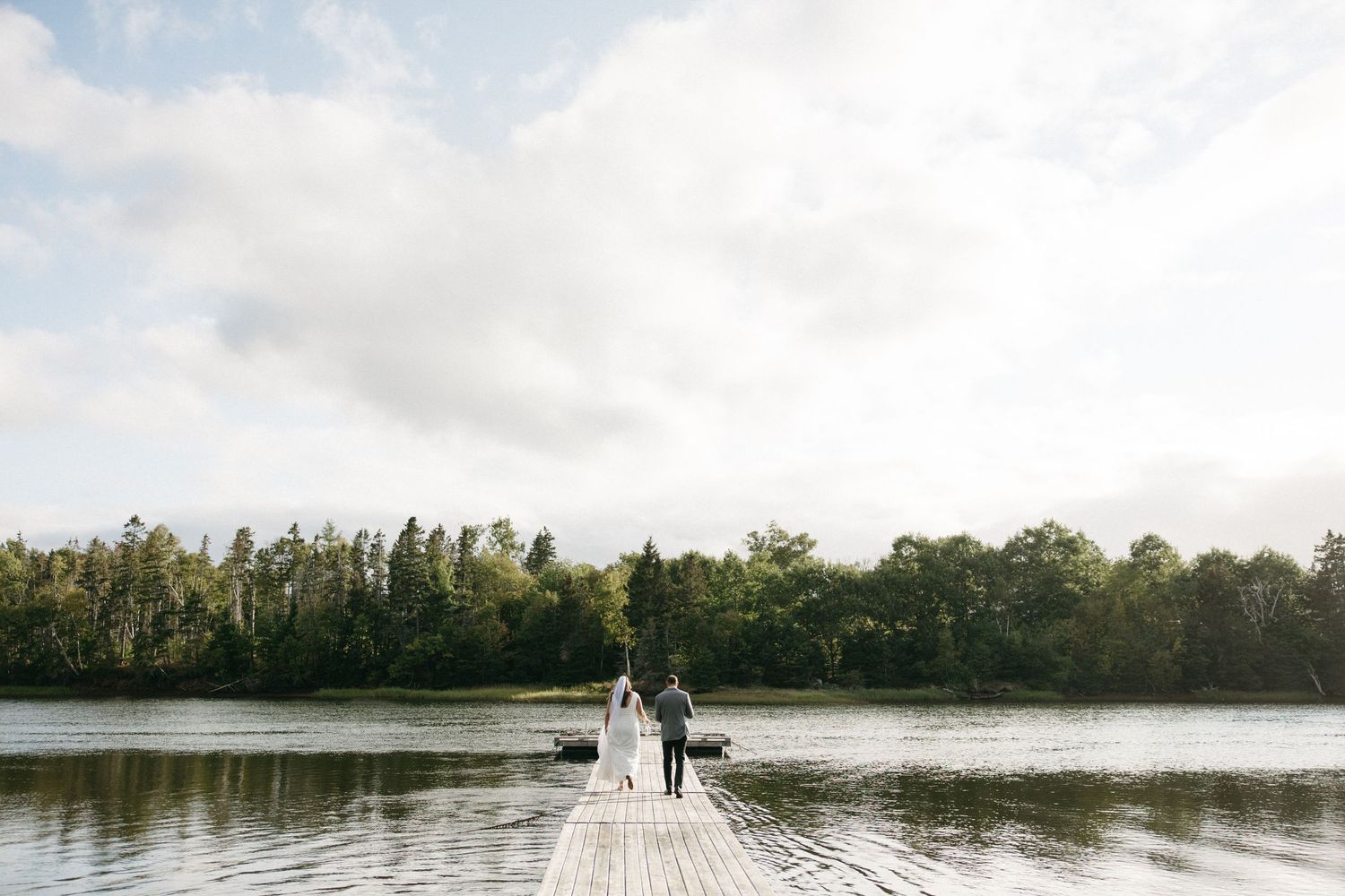 A wooden dock stretches out over a serene lake surrounded by trees on a cloudy day.