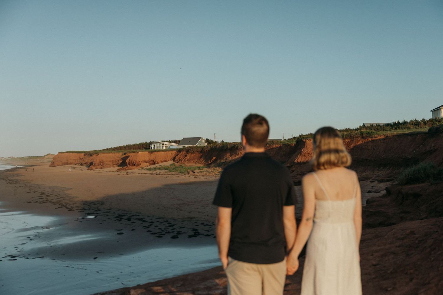 A couple walks along a scenic coastal beach with cliffs and ocean waves during sunset.