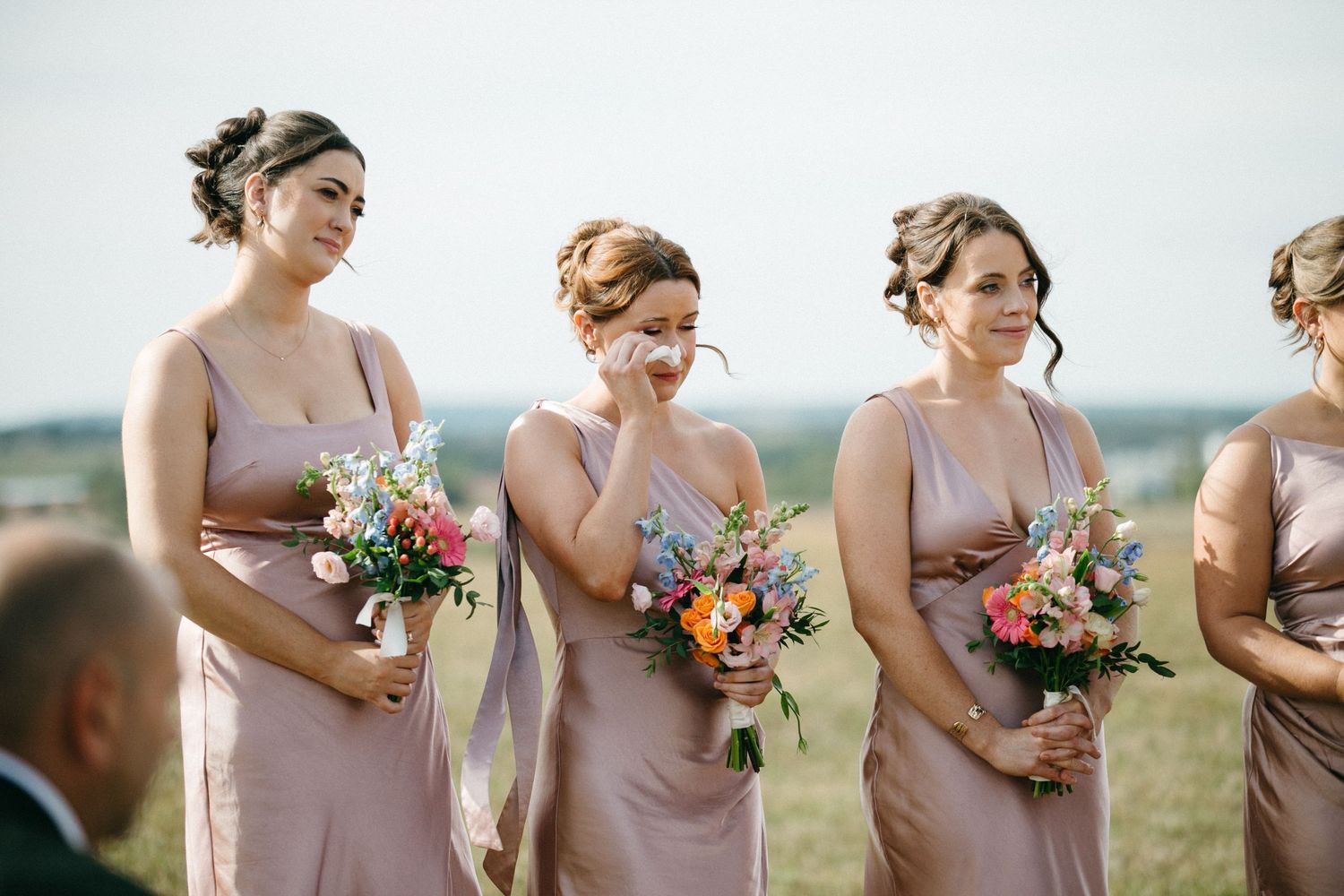 Bridesmaids in blush dresses hold bouquets while standing in a field.