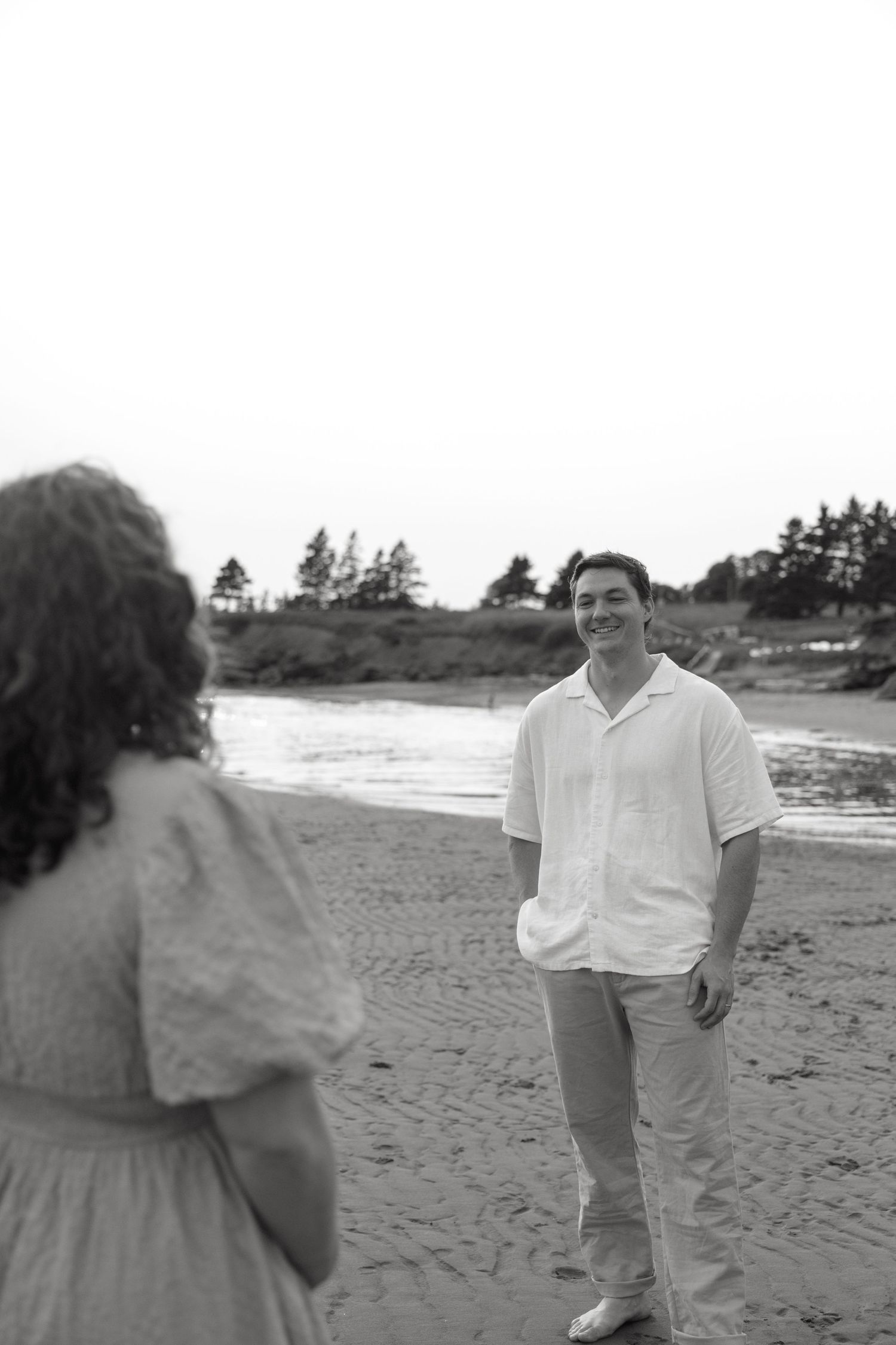 A couple enjoys a casual stroll together on a sandy beach with trees in the background.