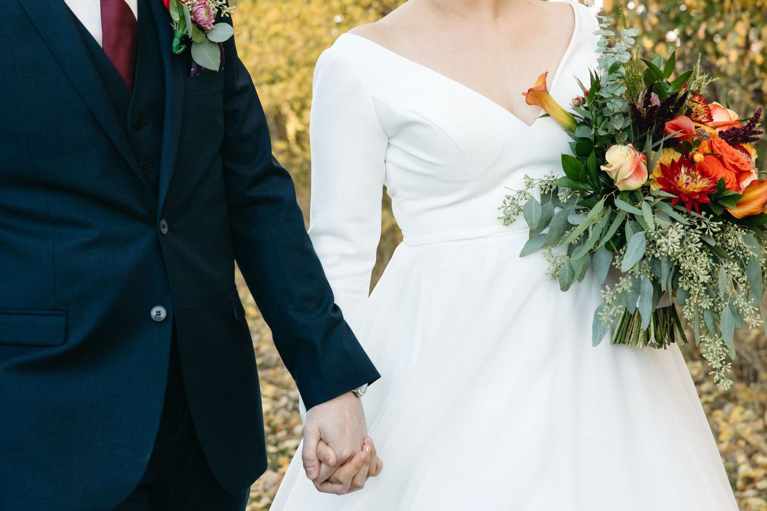 Wedding couple holding hands and walking together with autumn foliage and bridal bouquet.