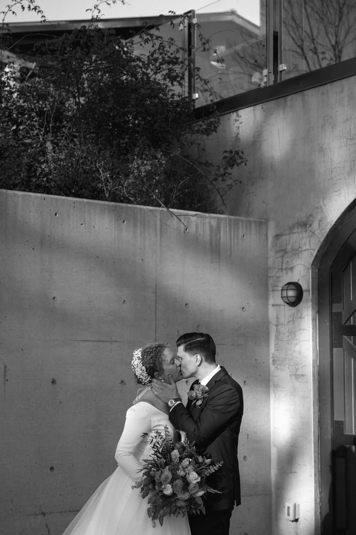 Black and white photo of a couple embracing by a concrete wall and arched doorway.