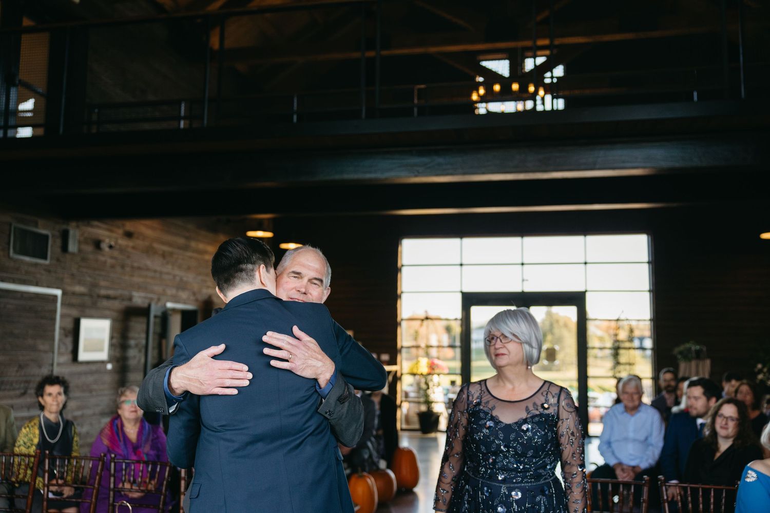 Wedding guests embrace and dance in a rustic venue with large windows.