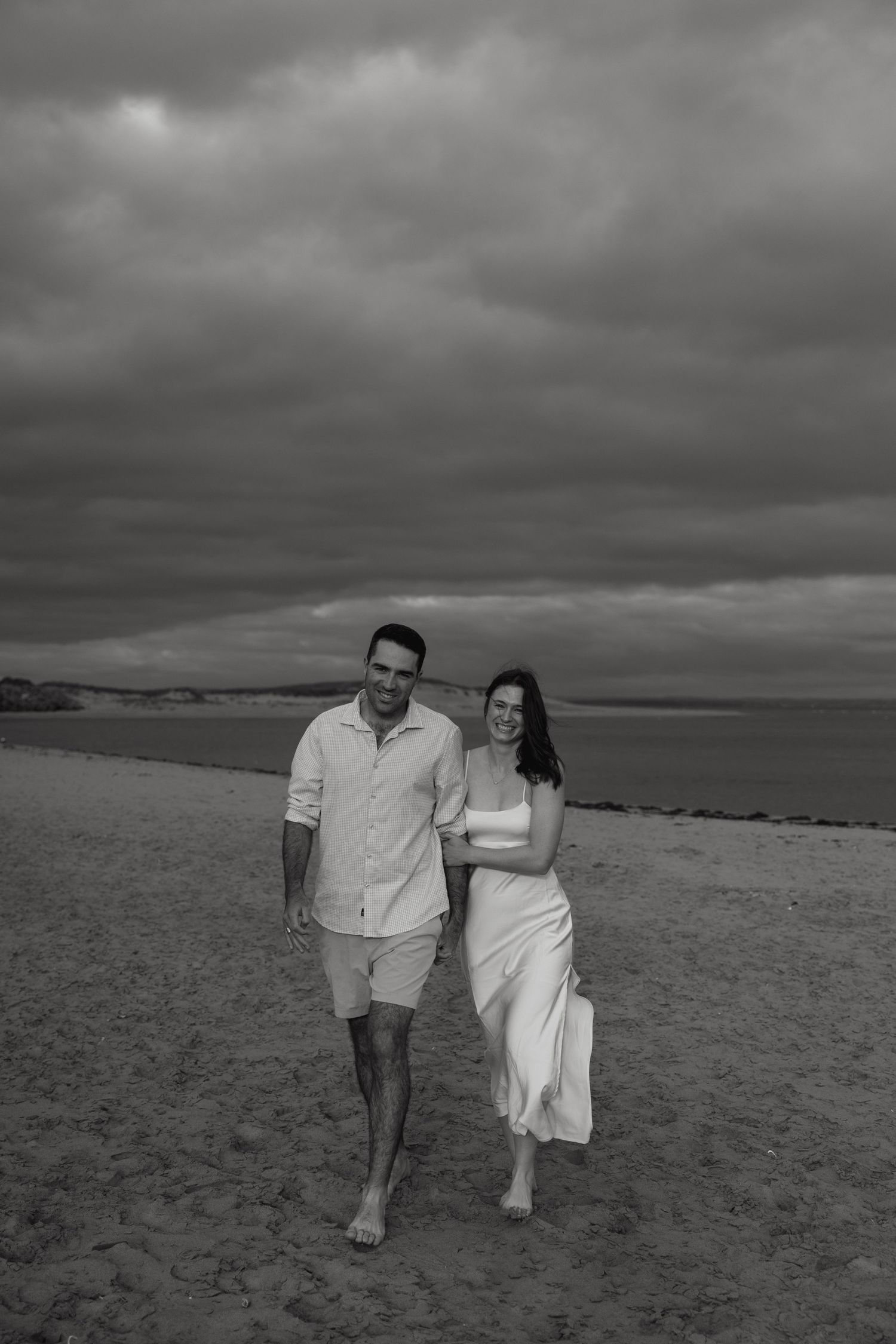 A couple walking together on a moody beach under dramatic stormy skies.