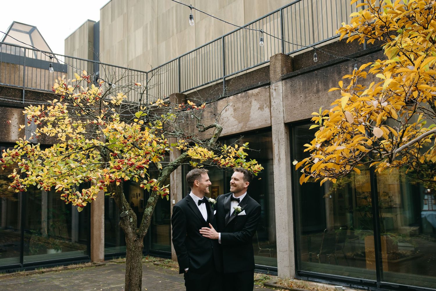 Two people in black tuxedos embrace under golden autumn trees in an urban setting.
