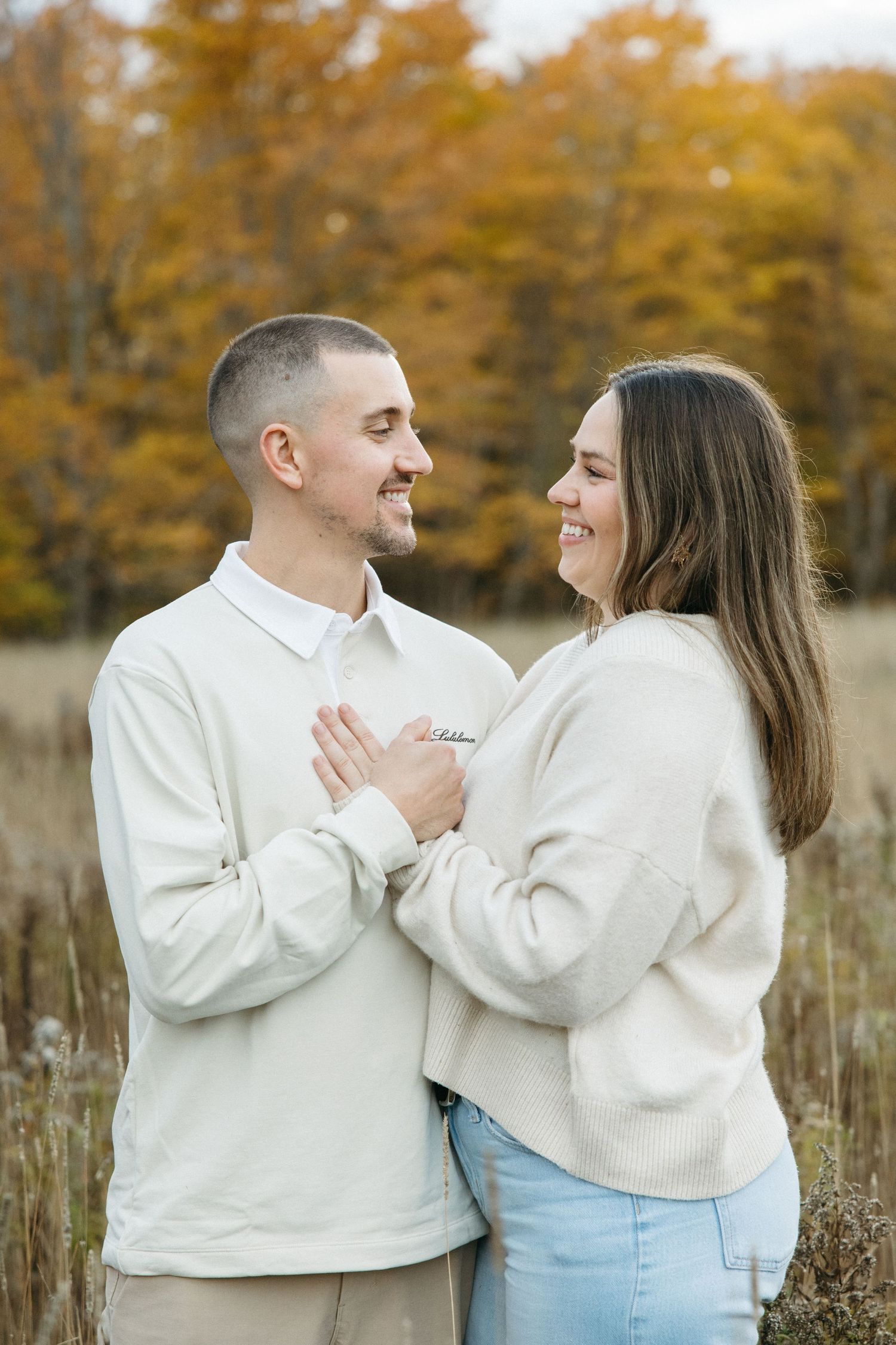 A couple in matching cream sweaters share a tender moment against an autumn backdrop.