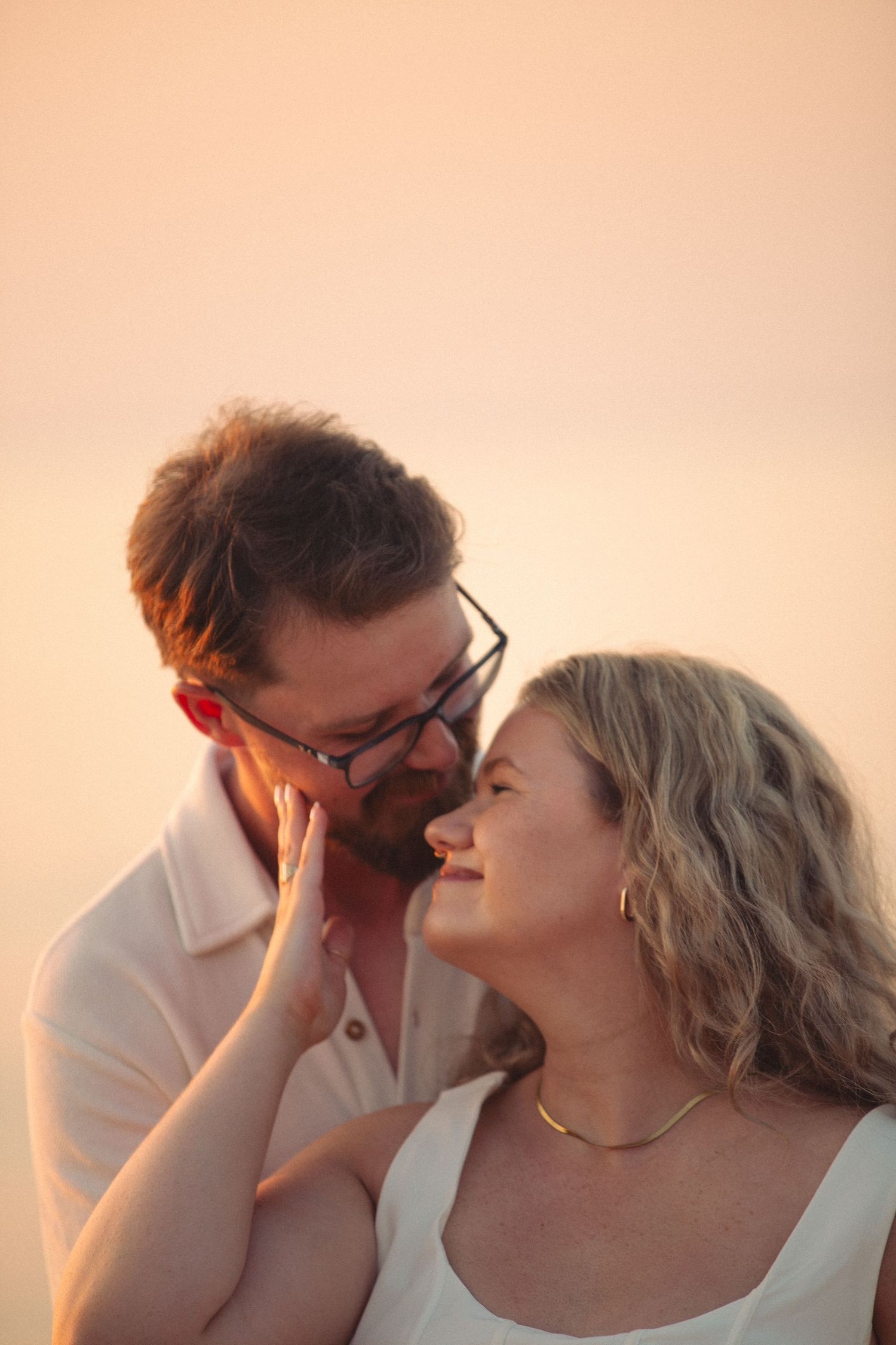 A couple shares a tender moment at golden hour, the man leaning in toward a smiling blonde woman wearing a gold necklace.