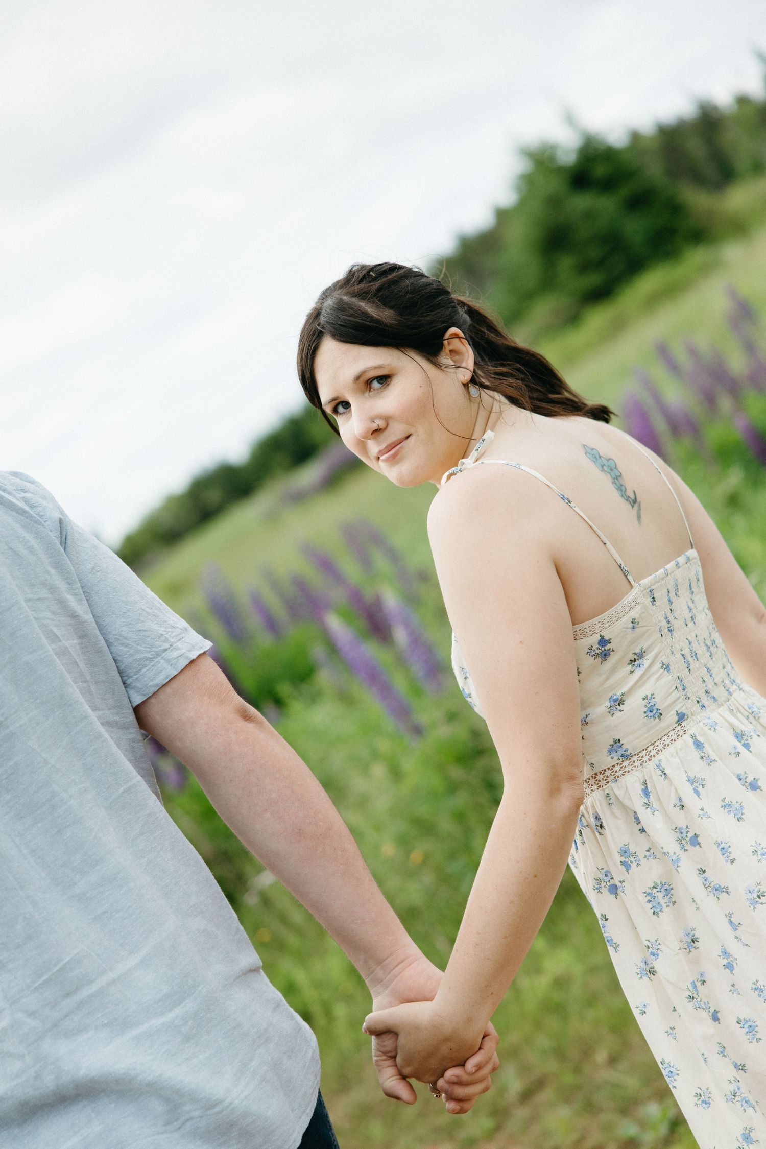 A young woman in a floral dress holds hands with a man while looking back, standing in a field of purple lupines.
