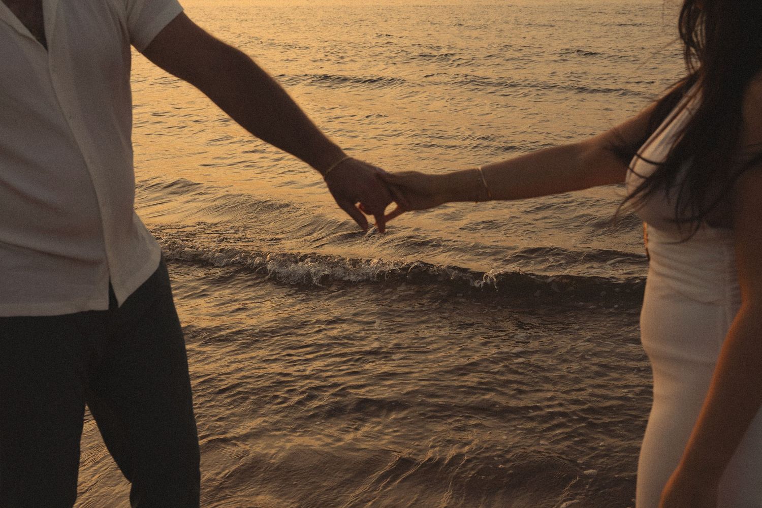 Romantic couple holding hands at ocean's edge during golden sunset, silhouetted against shimmering water.