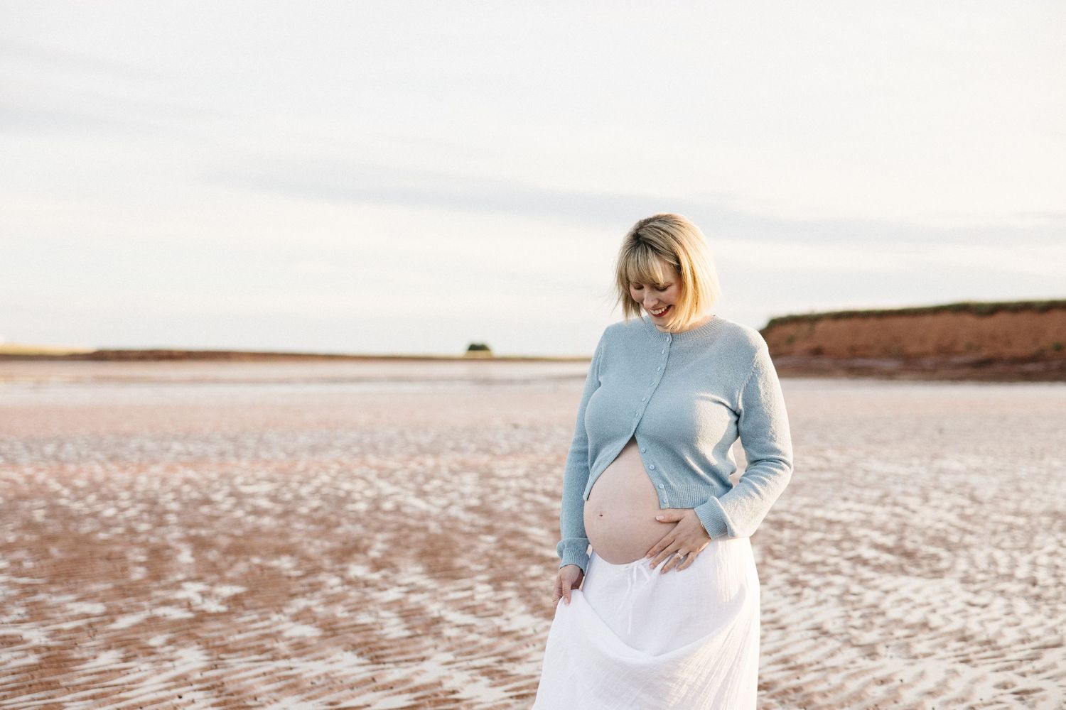 Pregnant woman in blue sweater and white skirt cradling baby bump outdoors on a sandy beach at sunset.