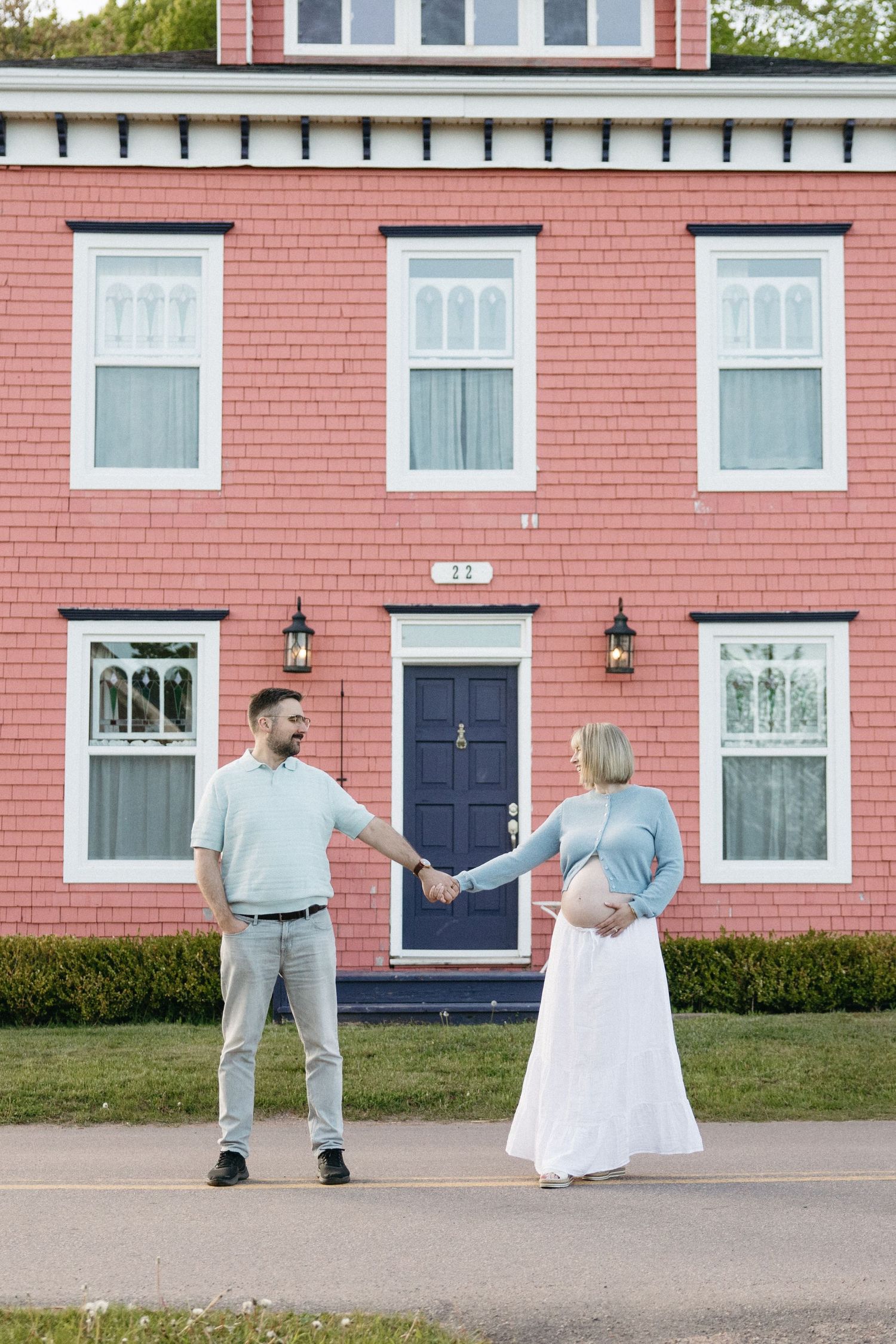 Pregnant couple holding hands in front of a red brick house with a navy blue door.