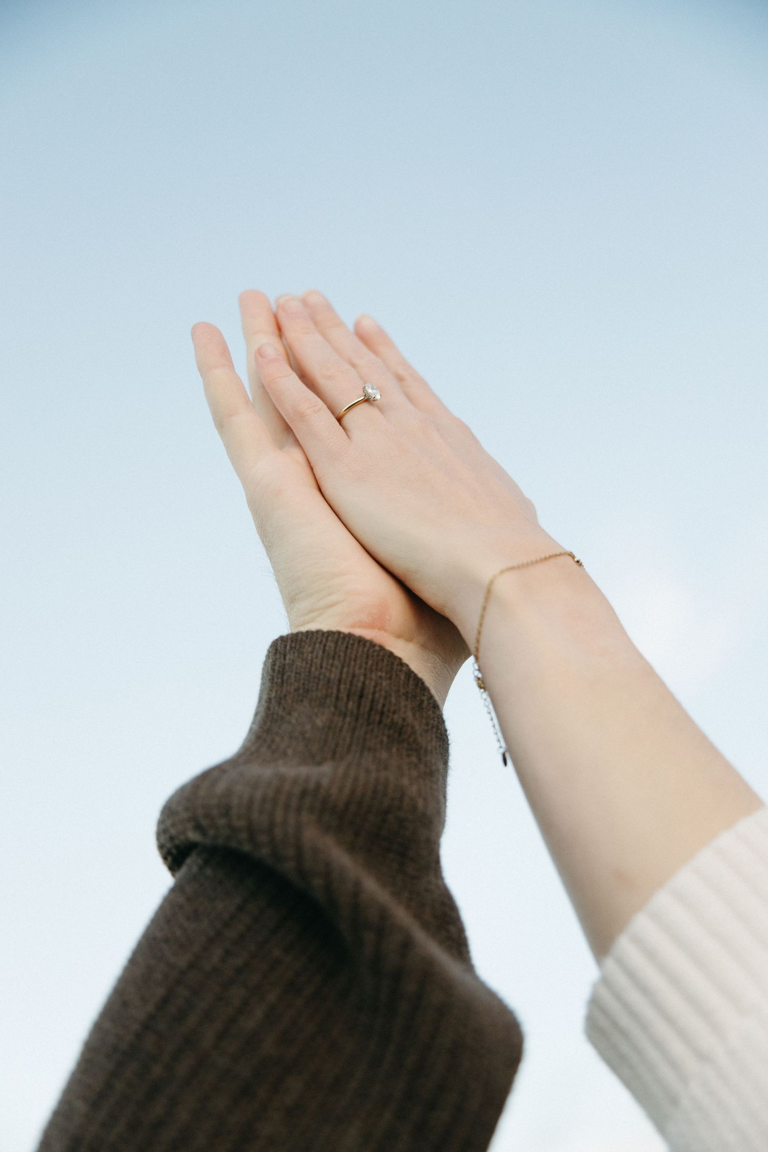Two hands reach toward a pale blue sky, one wearing a delicate diamond ring and a gold bracelet.