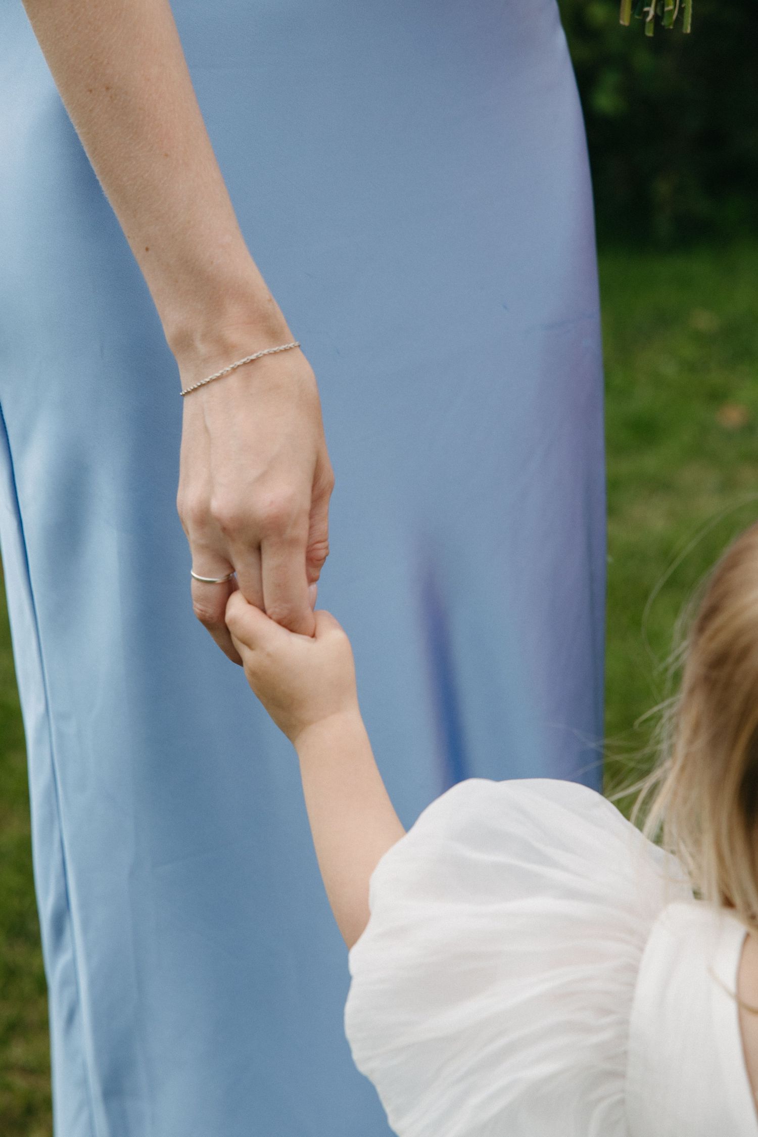 Woman in blue dress holding hands with young child in white dress outdoors.