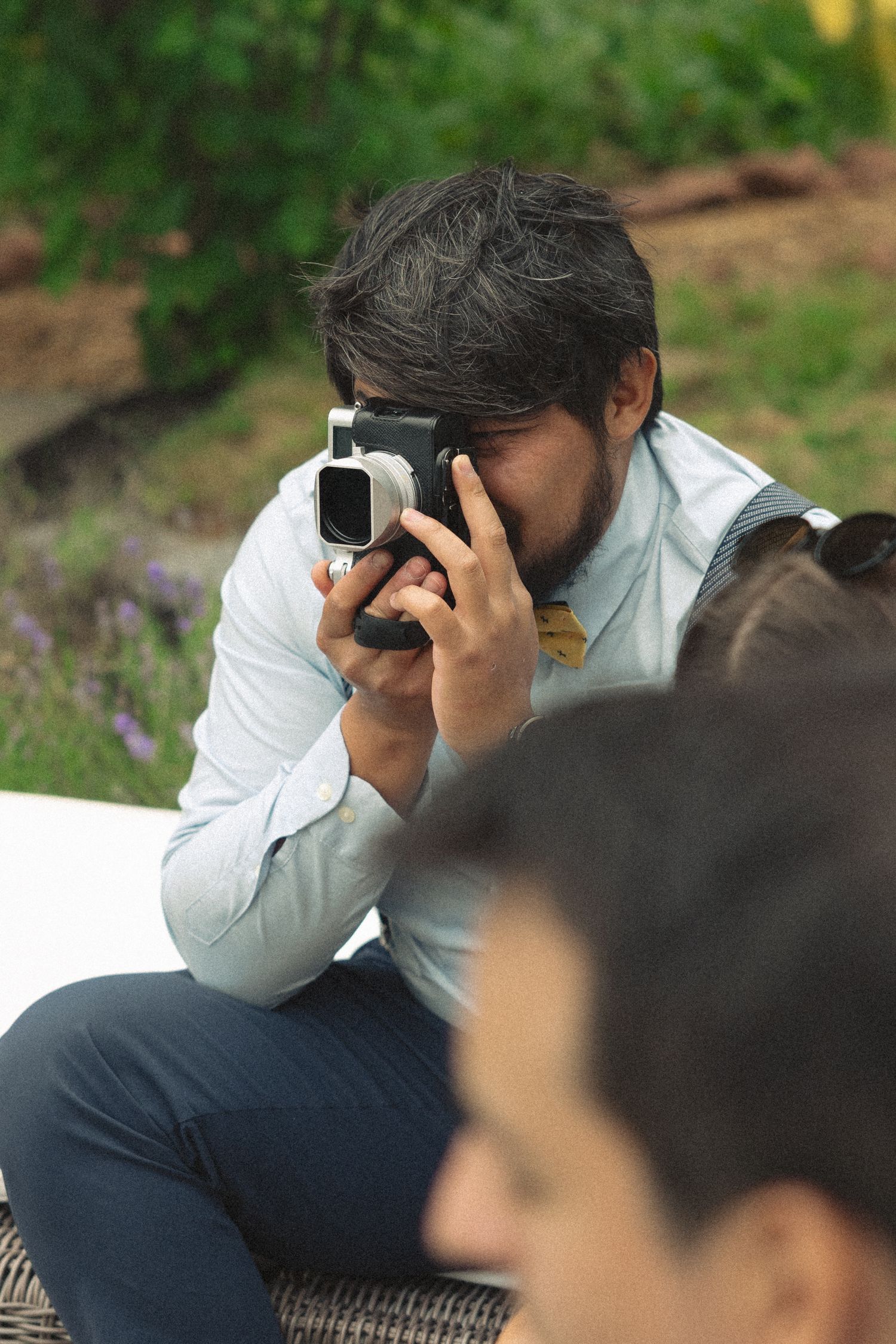 Bearded man in white shirt photographing with vintage camera at outdoor wedding reception.