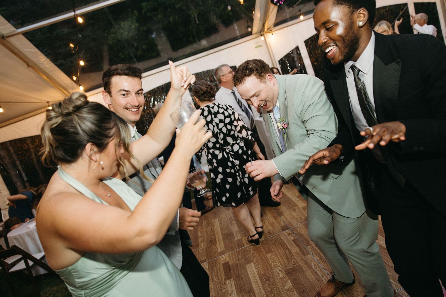 Wedding guests in mint and sage attire laugh and dance together under a clear tent at the reception.