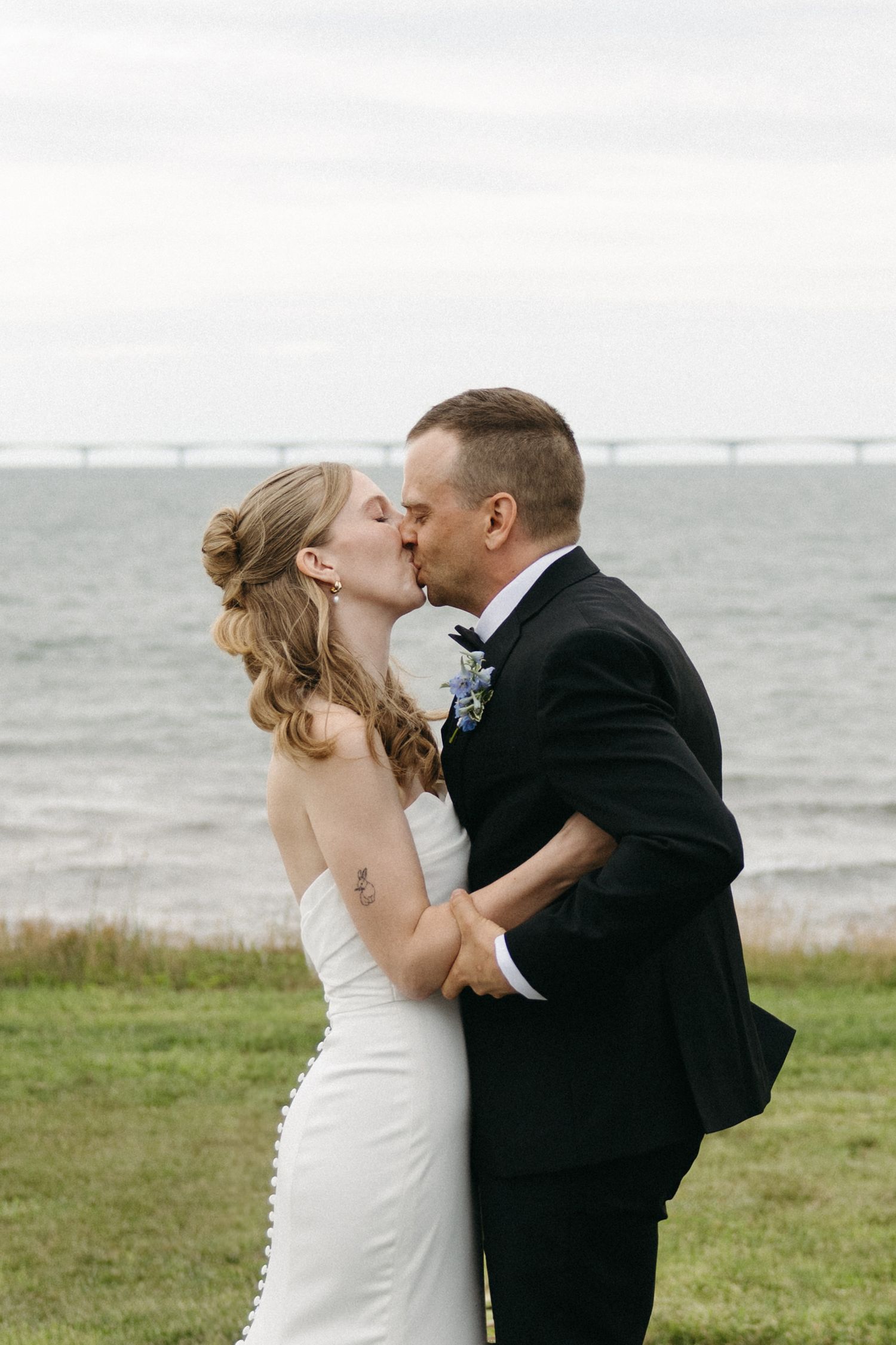 Bride and groom share romantic kiss by the waterfront with bridge visible in background.