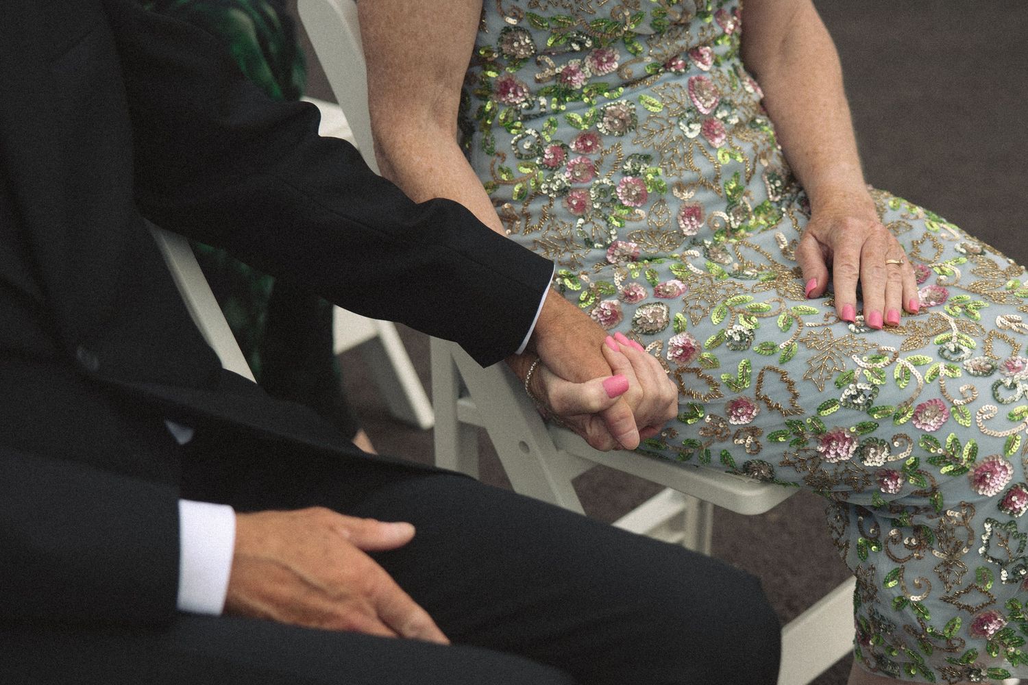 Couple holds hands tenderly during outdoor wedding ceremony, dressed in floral and formal attire.