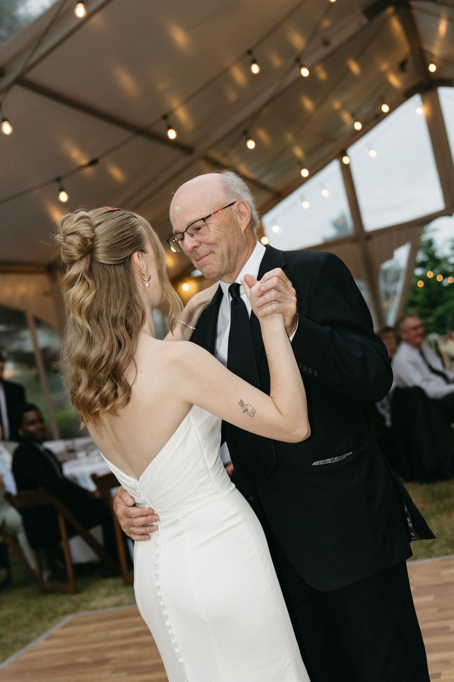 Bride in fitted white gown shares an emotional father-daughter dance under warm string lights in a clear tent.